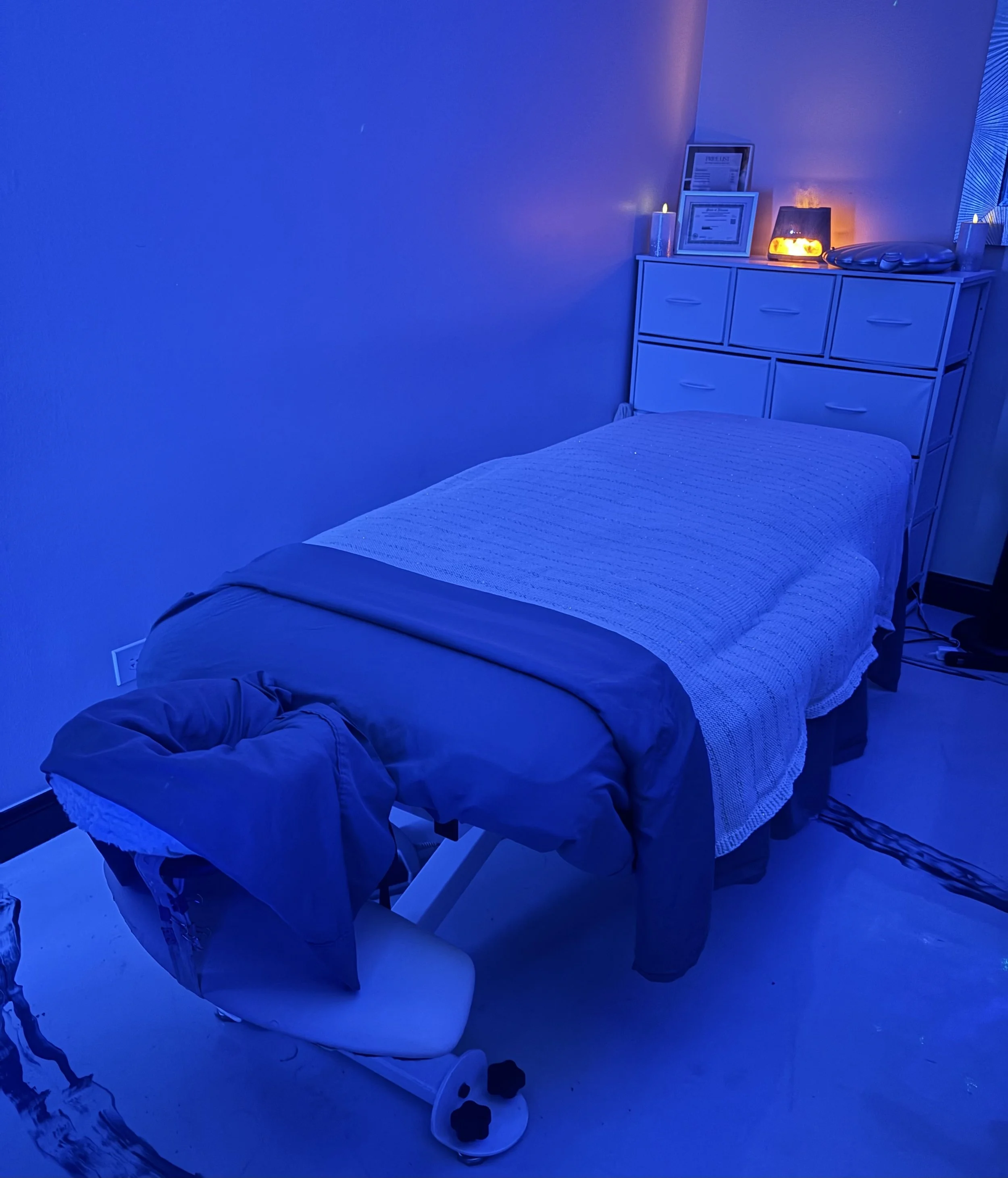 A cozy bedroom with a bed covered in a white textured blanket, a white dresser with framed photos, documents, candles, a diffuser, and a decorative lamp emitting warm light. The room is lit with calm blue lighting, creating a relaxing atmosphere.