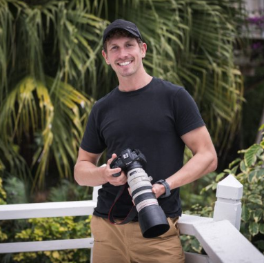 Man with a camera outdoors with lush green plants in the background.