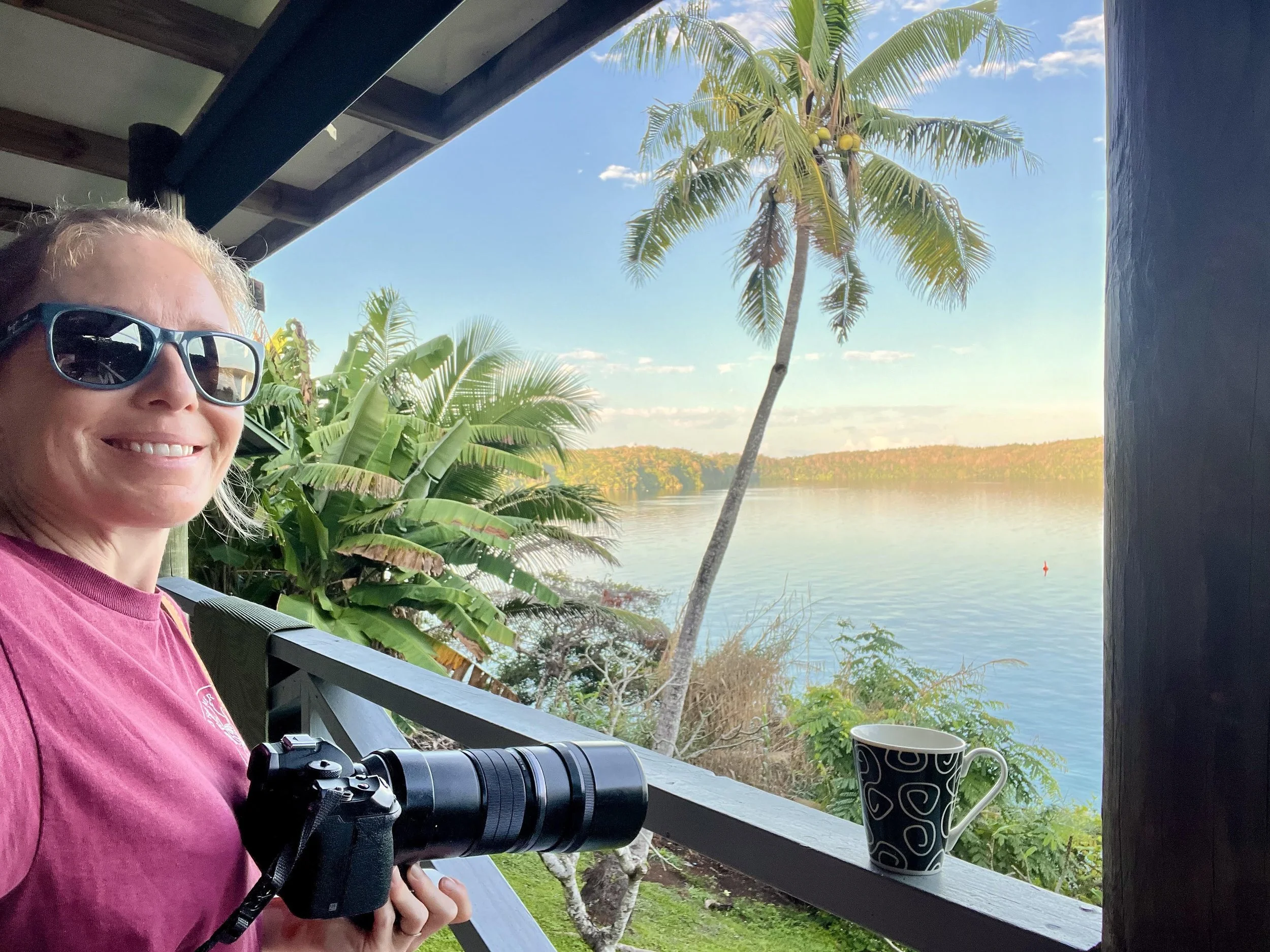 A smiling woman with blonde hair wearing sunglasses and a pink shirt, holding binoculars, is standing on a porch overlooking a river with green trees and a blue sky with some clouds. There is a black and white patterned mug on the porch railing.