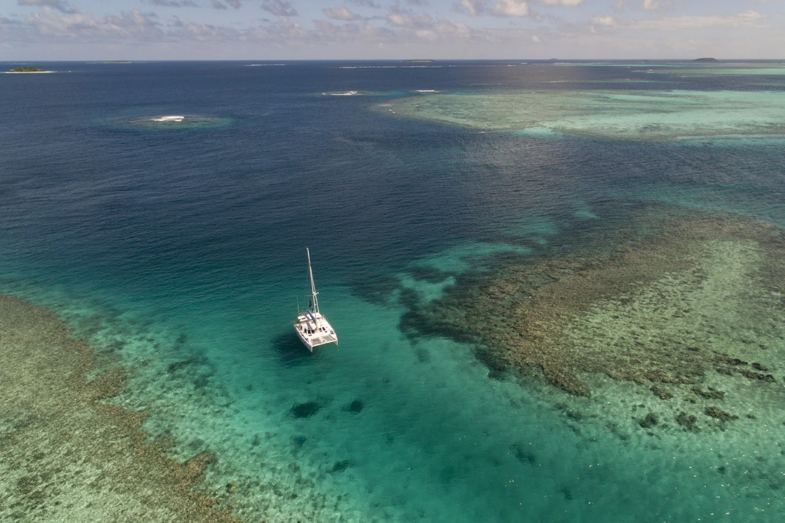 An aerial view of a sailboat anchored in clear turquoise waters near a coral reef with patches of seaweed and underwater formations, with small islands on the horizon under partly cloudy sky.