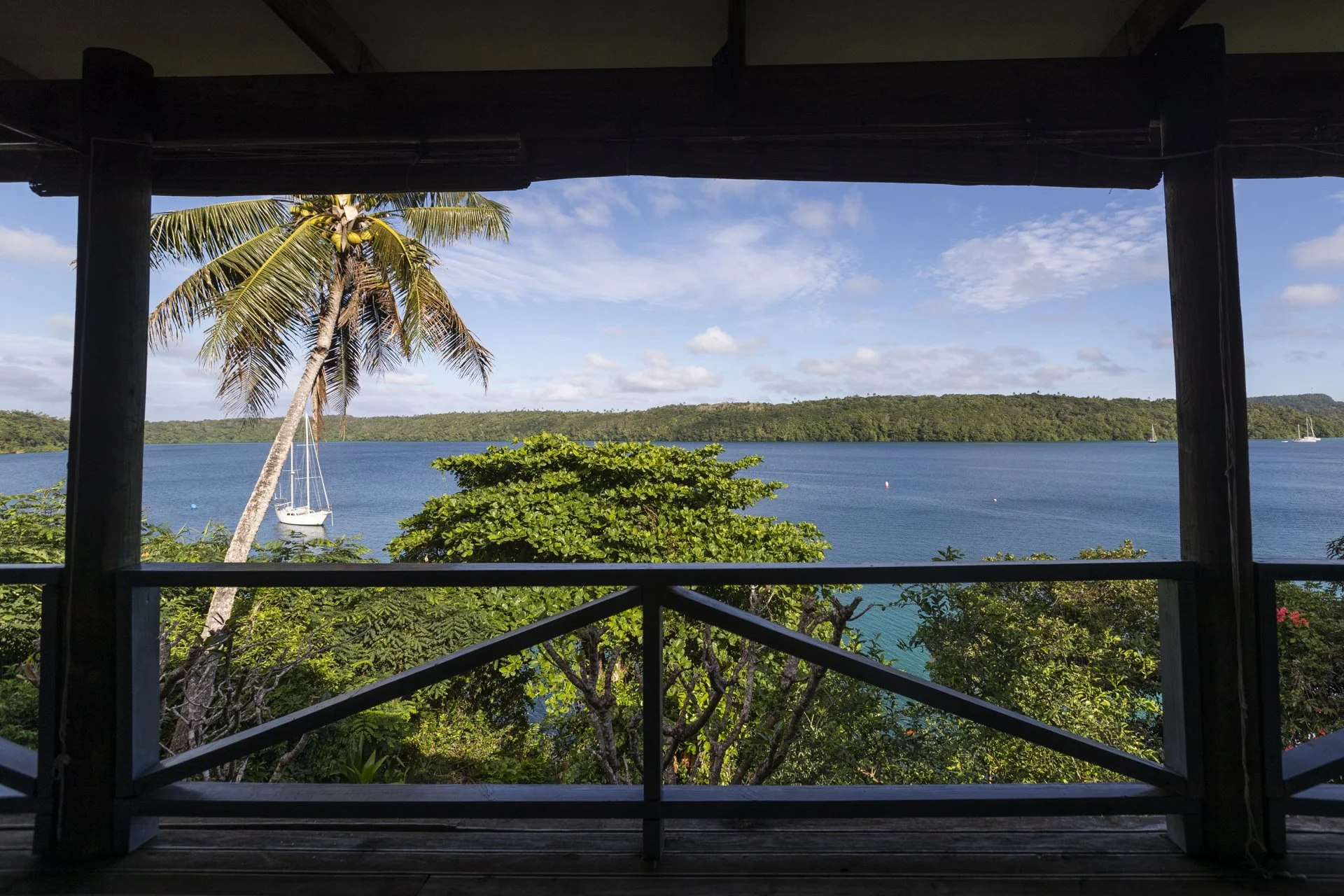 View of a tropical lake with a sailboat, surrounded by lush greenery and distant hills, seen from a balcony with wooden beams and railing.