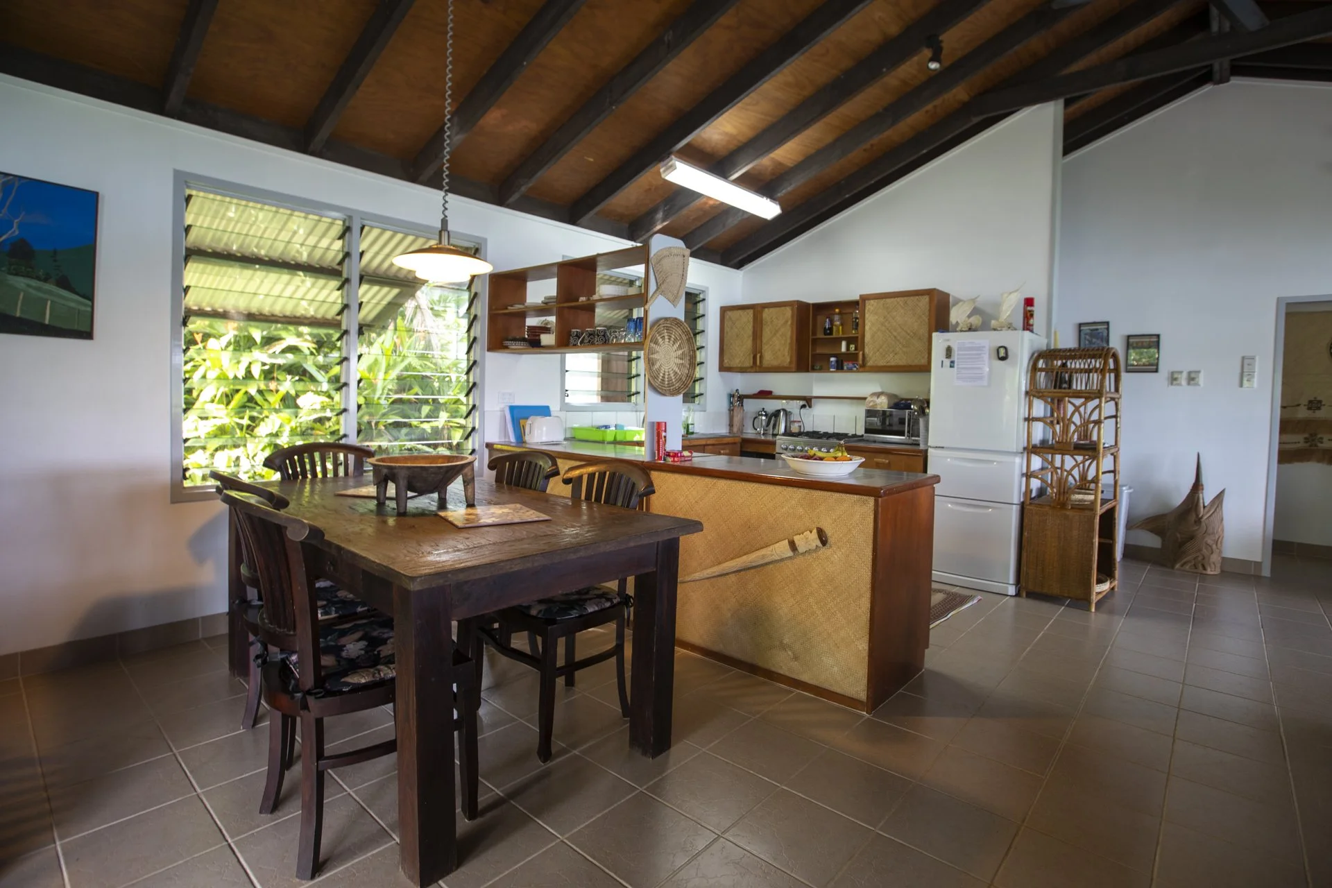 Interior view of a dining area and kitchen with wooden dining table and chairs, open shelving, a white refrigerator, and tropical plants visible through window louvers.