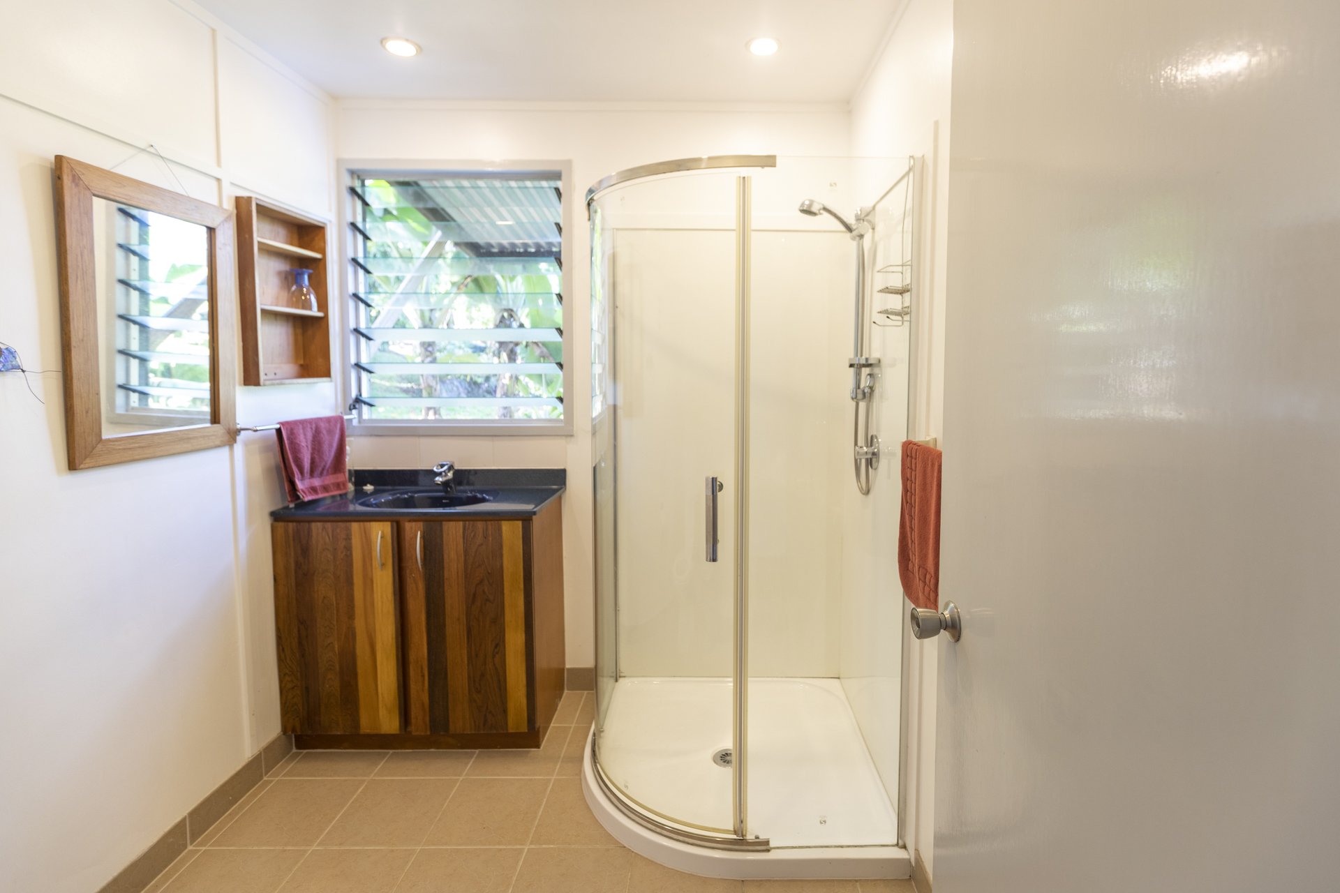 A small bathroom with a shower stall with curved glass doors, a wooden cabinet with a black countertop, a large window with green plants outside, and a wall mirror with a red towel hanging.