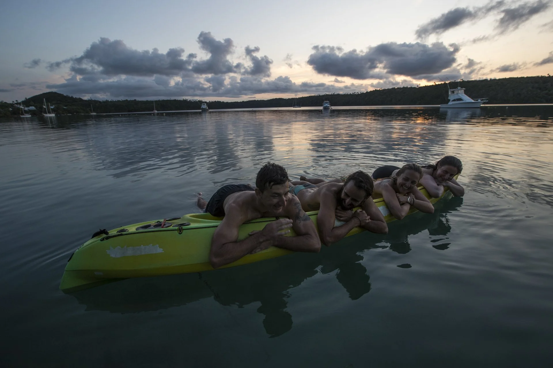 Four people holding onto a kayak in the water during sunset, smiling, with boats and a hilly shoreline in the background.