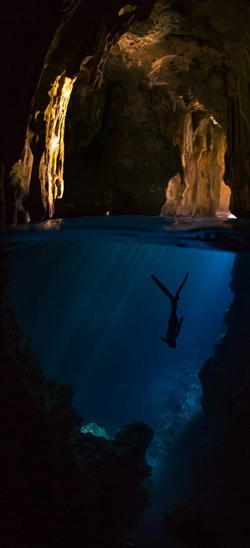Person swimming upside down in a cave with sunlight streaming through the opening, illuminating the water.