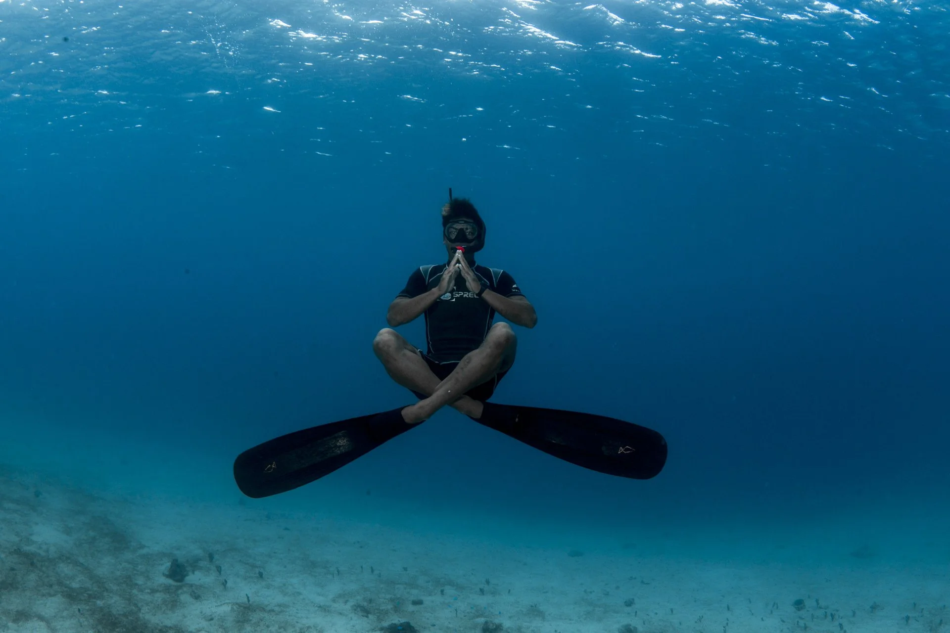 Male scuba diver sitting cross-legged underwater with hands in a prayer position, surrounded by clear blue water and a sandy ocean floor.