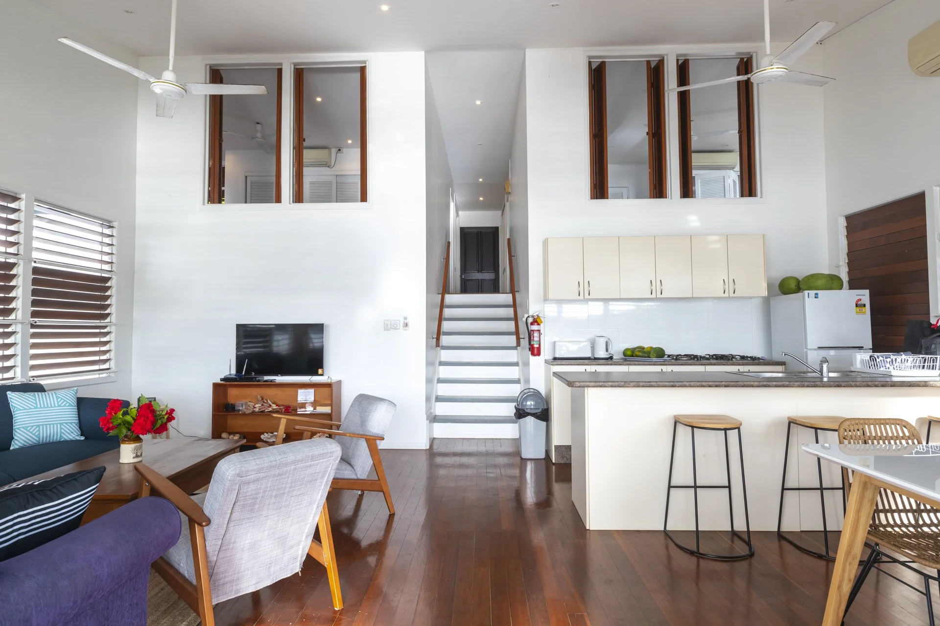 Open-concept living room and kitchen area with wooden floors, white walls, ceiling fans, and a staircase leading to an upper floor. The living area has a dark sofa with blue cushions, a wooden coffee table with a vase of red flowers, and various chairs. The kitchen has white cabinets, a countertop with green fruits, and bar stools. There is also a microwave, a toaster, and a refrigerator.