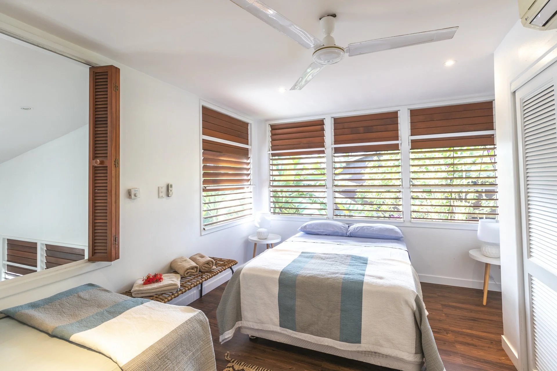 A bedroom with two beds, each covered with a beige quilt featuring blue and green stripes, located in front of large windows with wooden blinds. There are two small bedside tables with white lamps, a wooden bench with towels, and a ceiling fan, all in a bright room with white walls and wooden flooring.