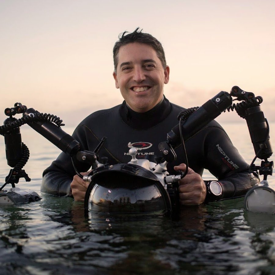 Man in wetsuit holding underwater camera equipment while in the water with sunset sky in the background.