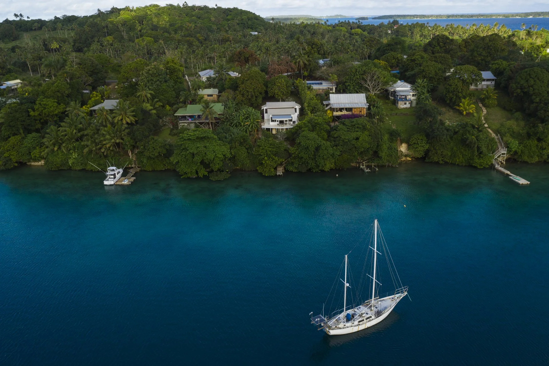Aerial view of a tropical waterfront with lush green trees, several houses, and a sailboat anchored in the calm blue water.