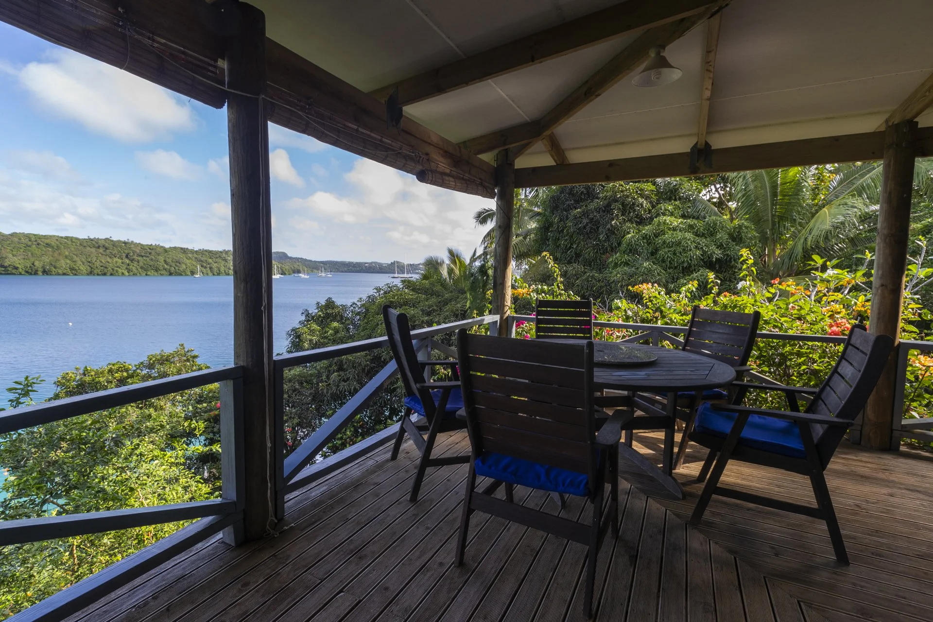 A wooden deck overlooking a body of water with trees and sailboats in the distance, partially shaded by a roof, with a round table and several chairs with blue cushions.