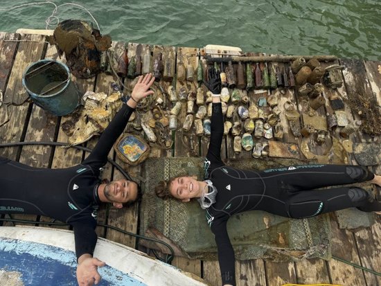 Two scuba divers lying on a wooden dock next to a collection of coral, shells, and marine debris washed ashore.