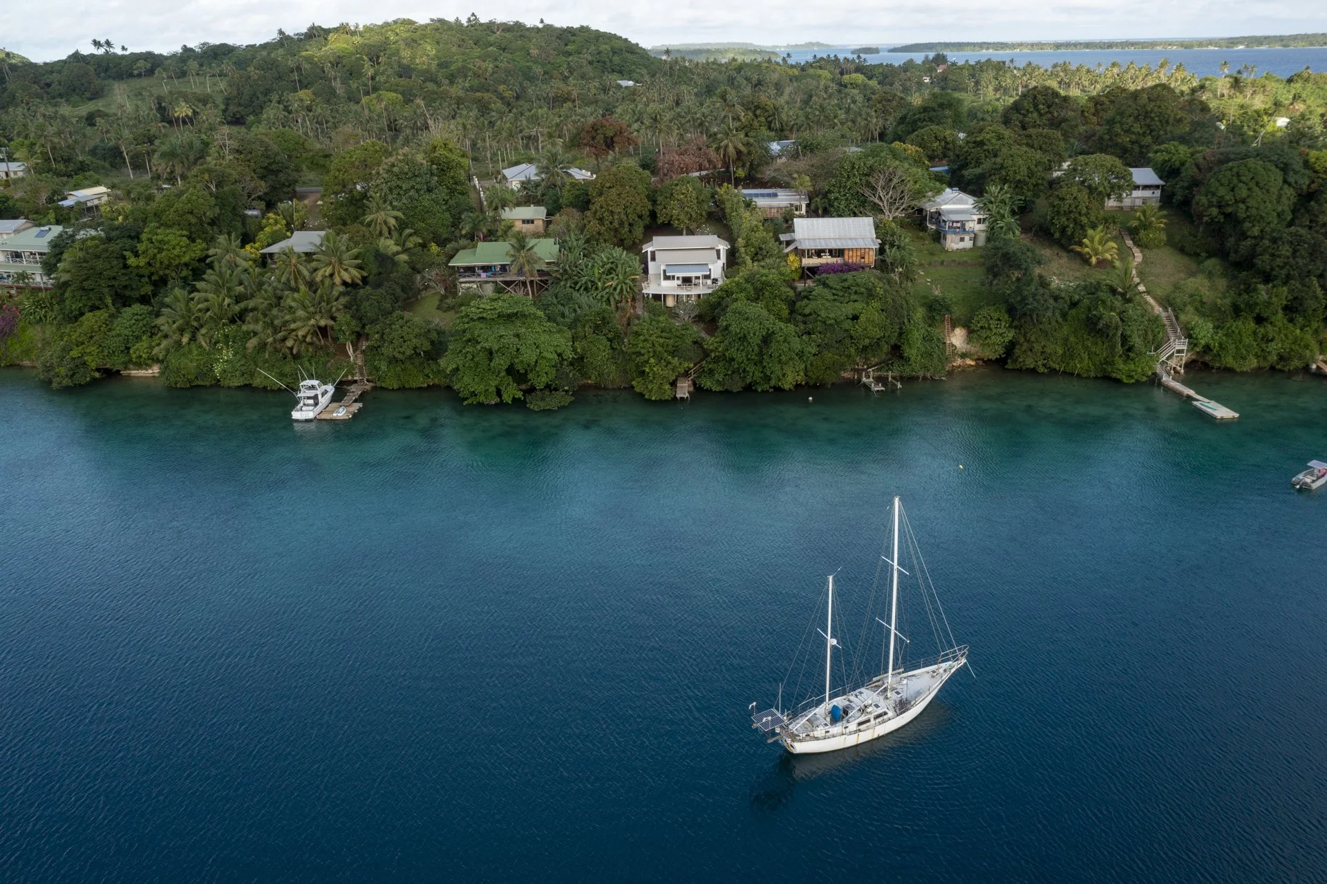 Aerial view of a coastal area with green trees, houses, and a dock, featuring a sailboat anchored in the blue water.