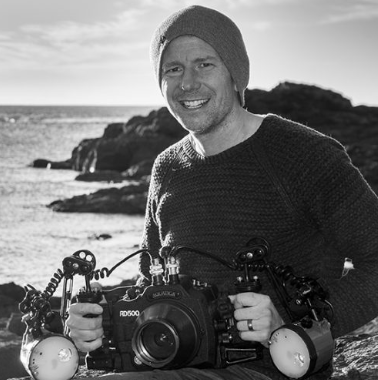Man smiling at the beach, holding a camera, wearing a knit sweater and beanie.