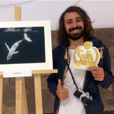 A man with curly hair and a beard is smiling and holding a gold medal with the text '1st' and 'GEA LA JOVEN' printed on it. He is standing next to an artwork of a whale swimming, mounted on an easel.
