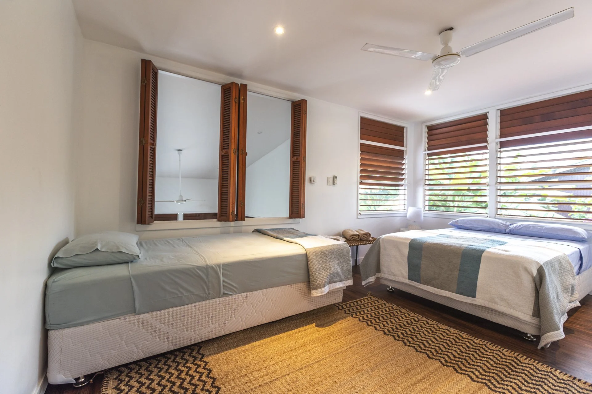 Bedroom with two beds, one with a mattress and the other with a quilted bedspread, wooden shutters on the windows, mirror on the wall, ceiling fan, and a small woven rug on the floor.