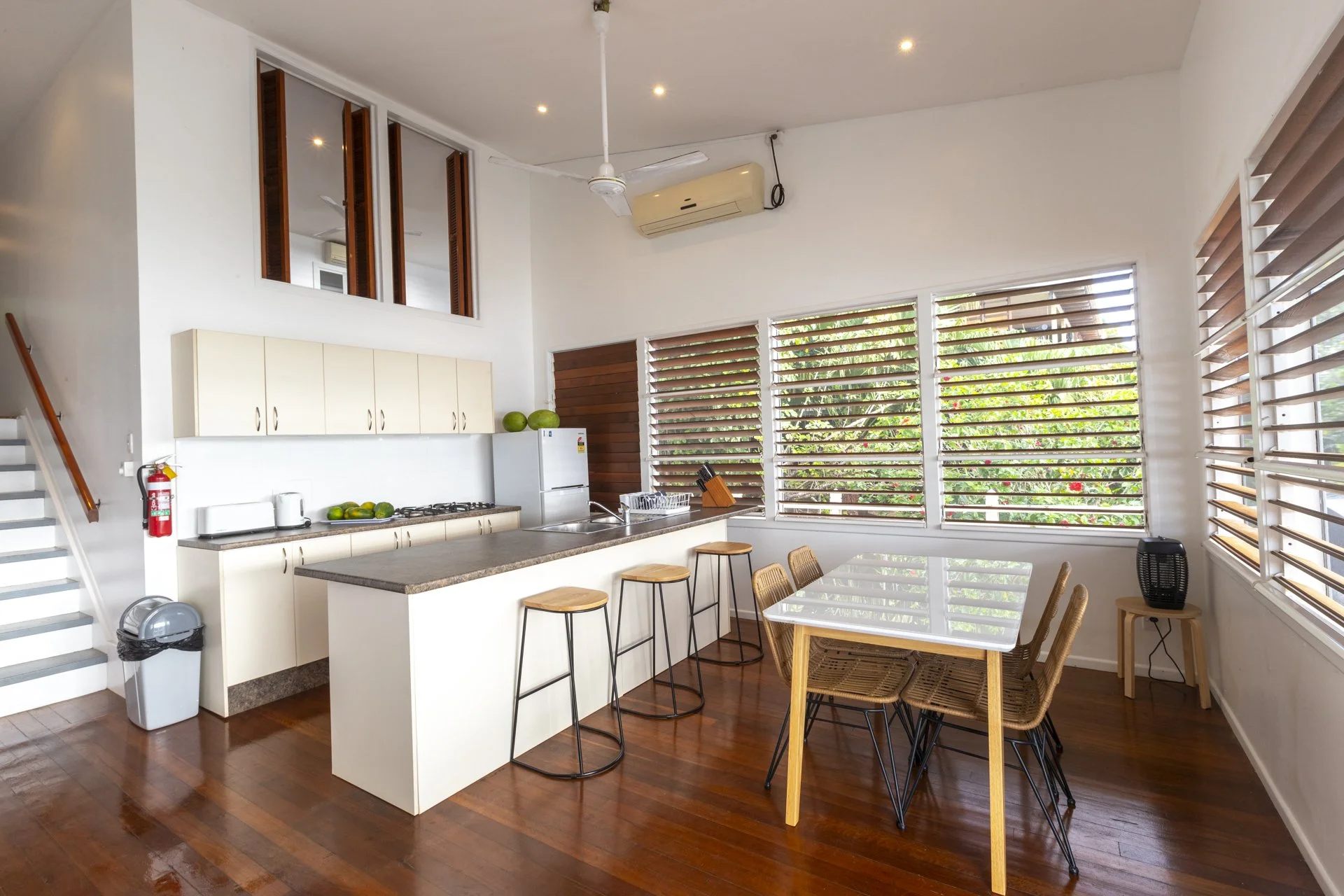 Bright kitchen with large windows, white cabinets, a countertop, a small dining table with wicker chairs, and decorative green watermelons.