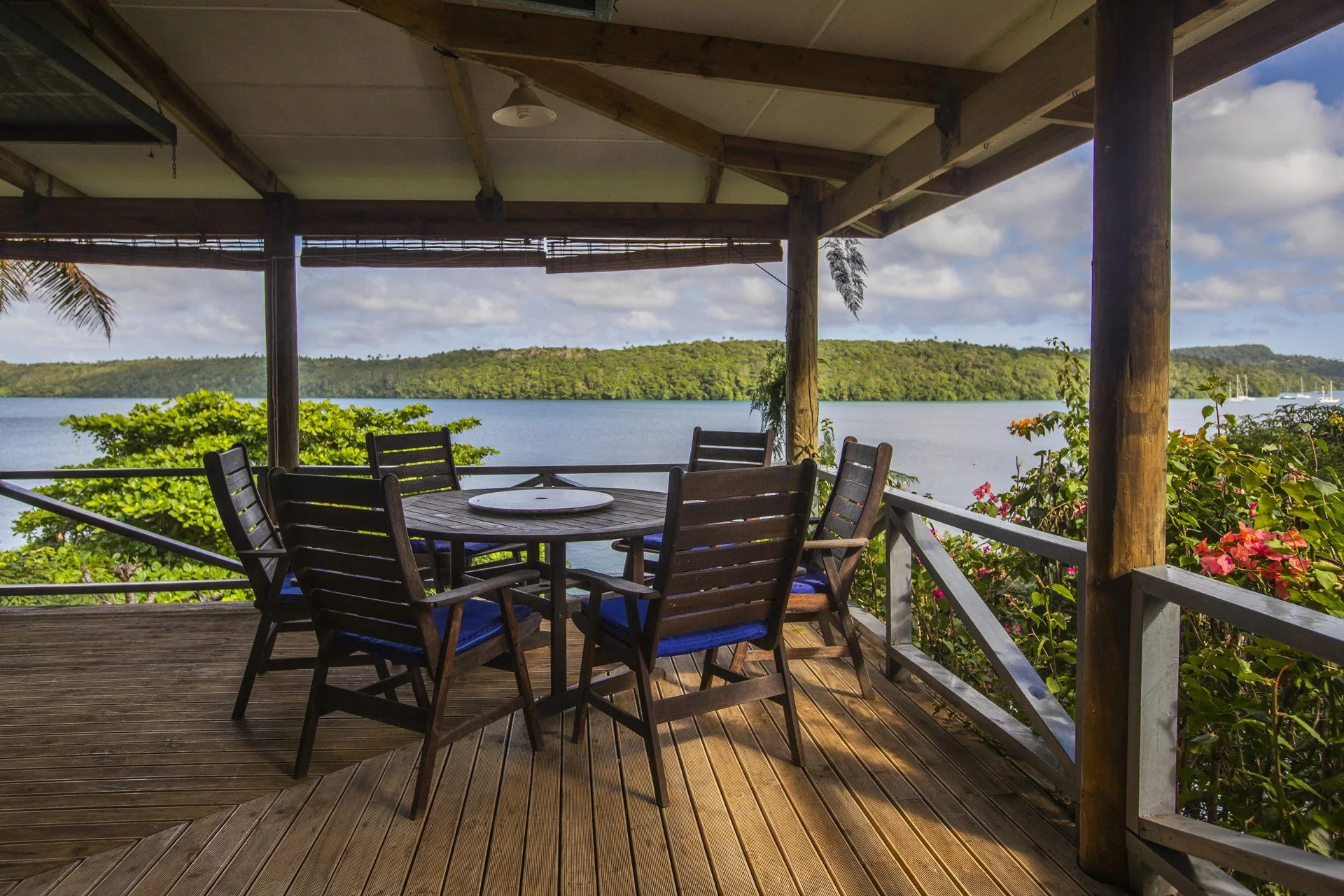 A wooden balcony with an outdoor dining set overlooking a body of water with lush green hills and a few sailboats in the distance