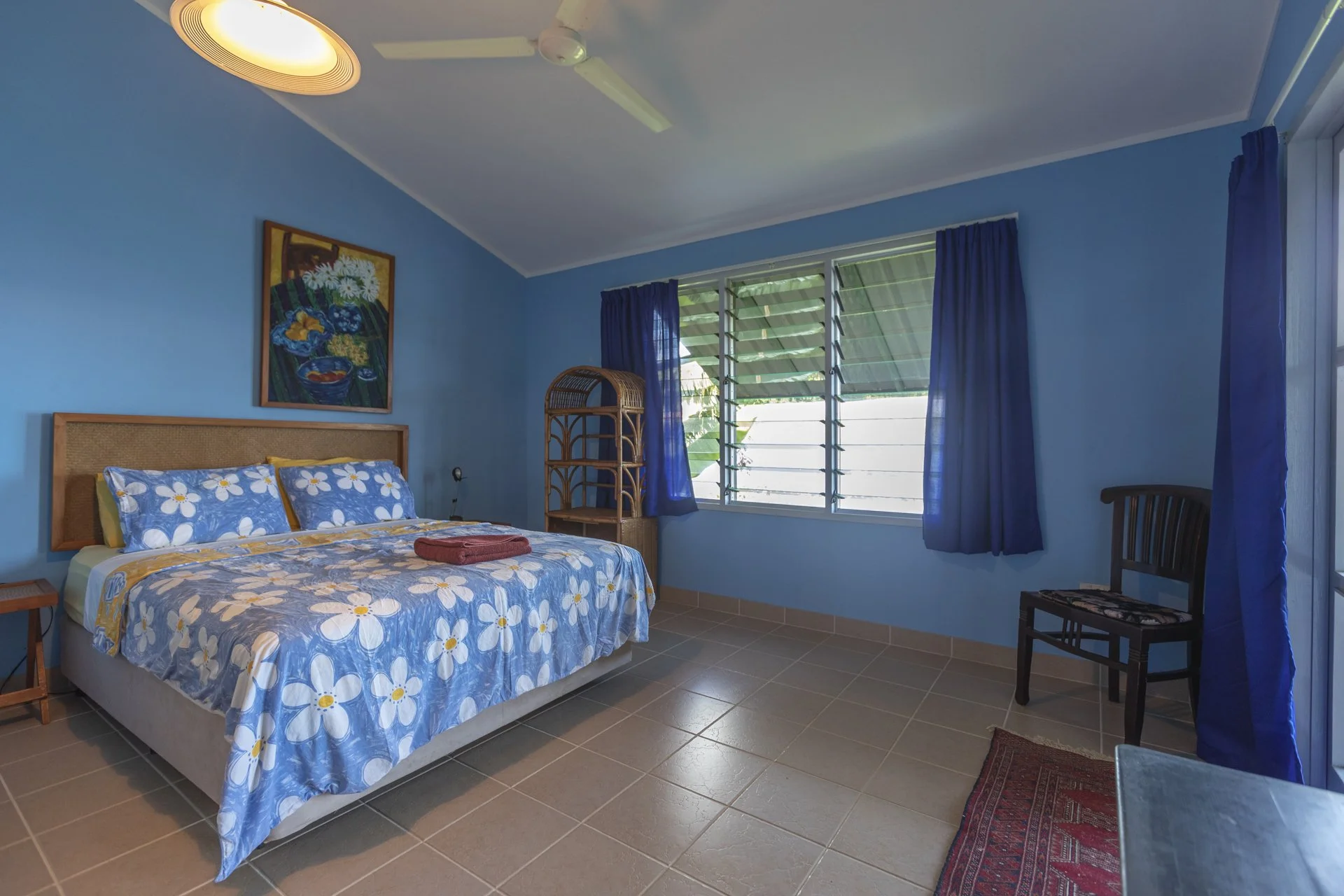 A bedroom with blue walls and curtains, a wooden bed with floral bedding, a wicker shelf, a chair, and a window with horizontal blinds.