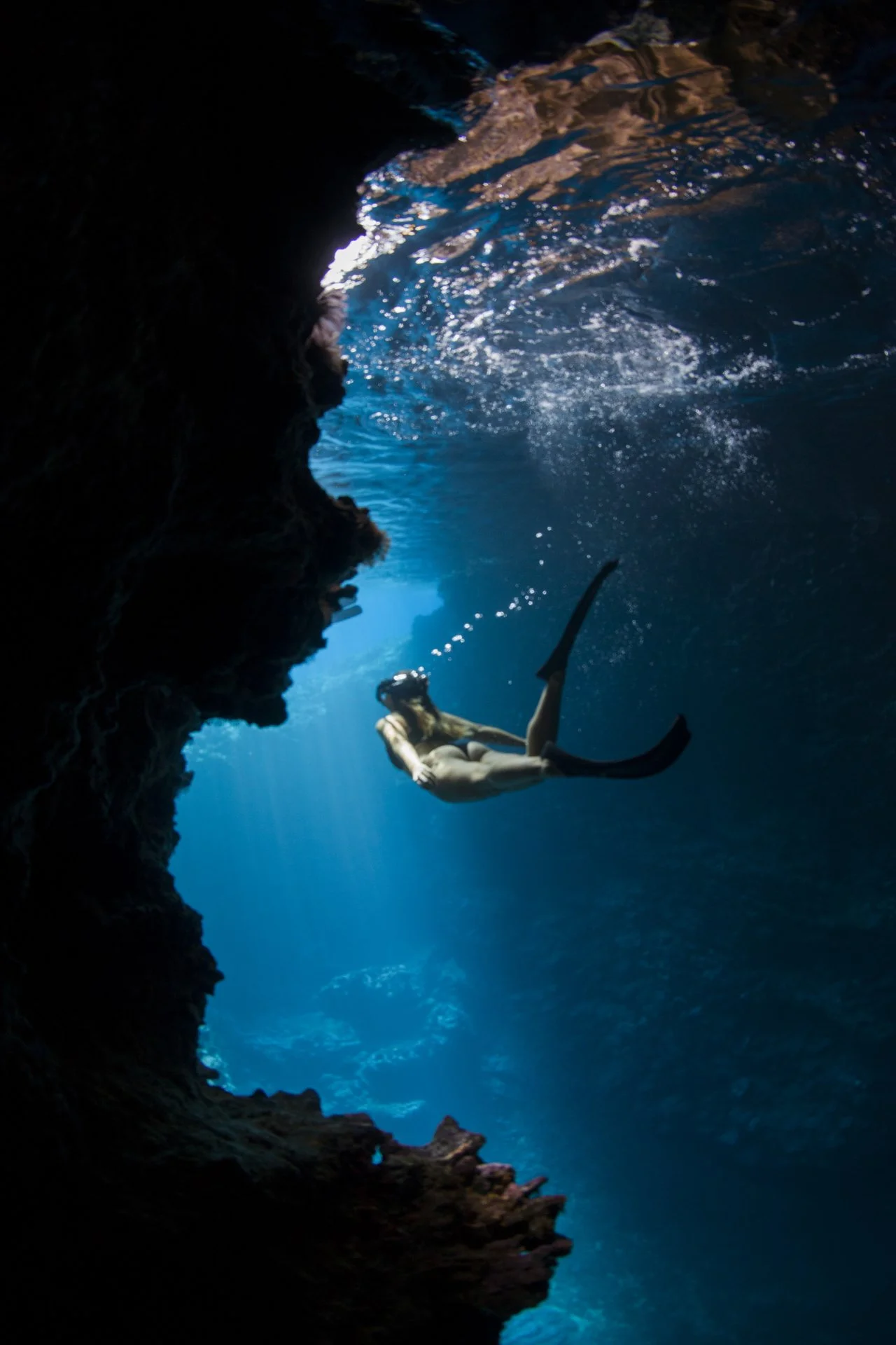 A person with long hair snorkeling underwater near a rocky cave passage, with sunlight illuminating the water and rocks.