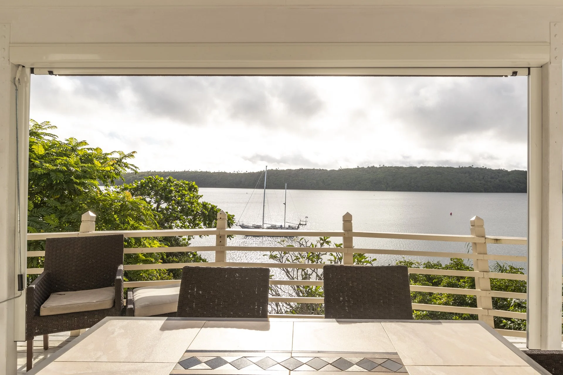 View of a boat on calm water with a forested hillside in the distance, seen through a large open window of a porch with outdoor furniture and a white railing.