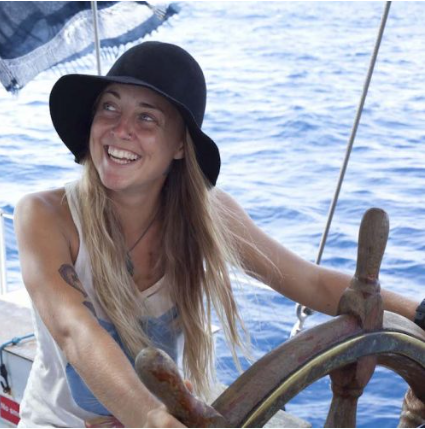 Woman with long blonde hair wearing a black hat, smiling while steering a boat over the ocean.