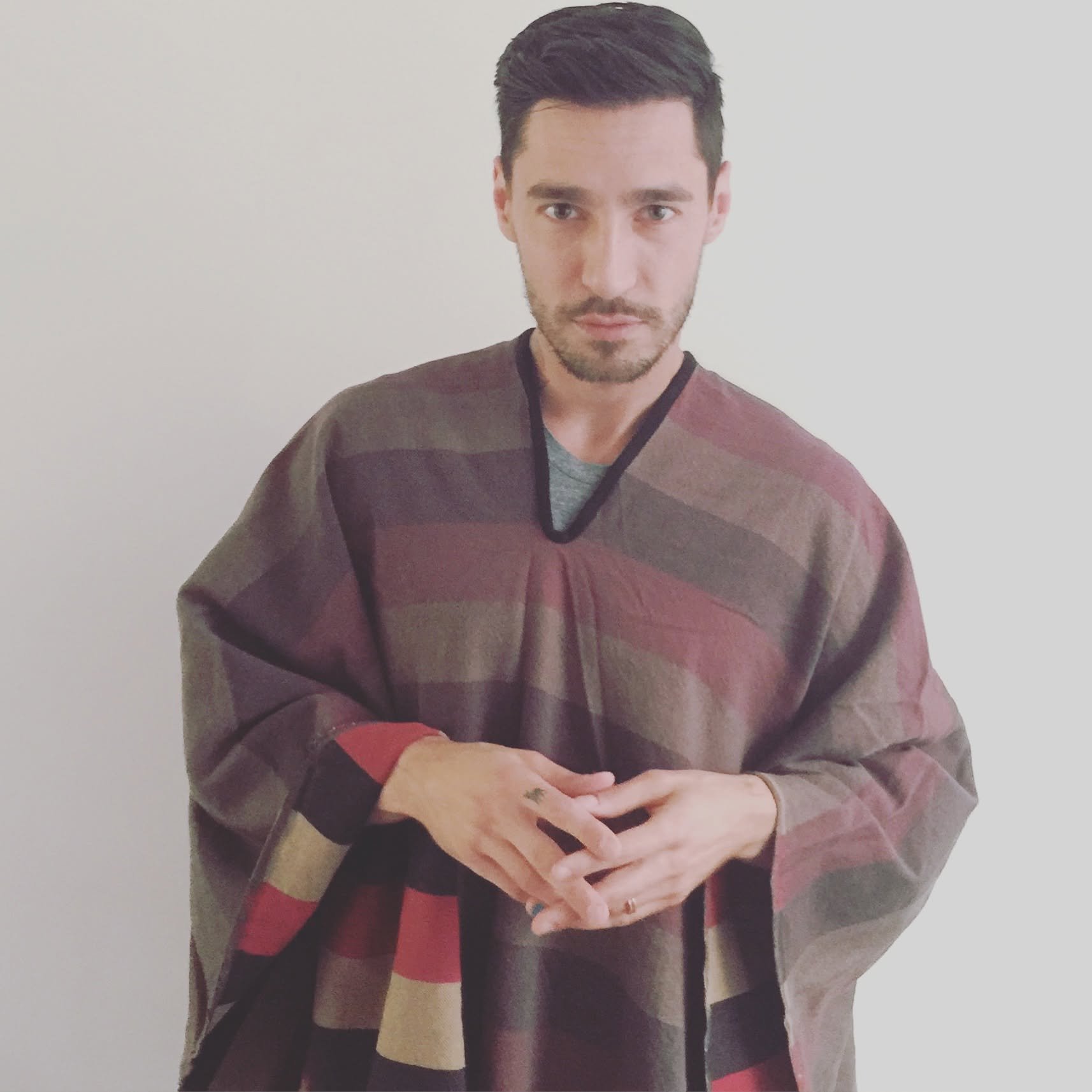 A man with dark hair, a beard, and light skin stands against a plain light-colored wall. He is wearing a dark-colored sweater with red and brown stripes and has a lanyard around his neck. His hands are positioned in front of him, with fingers intertwined.
