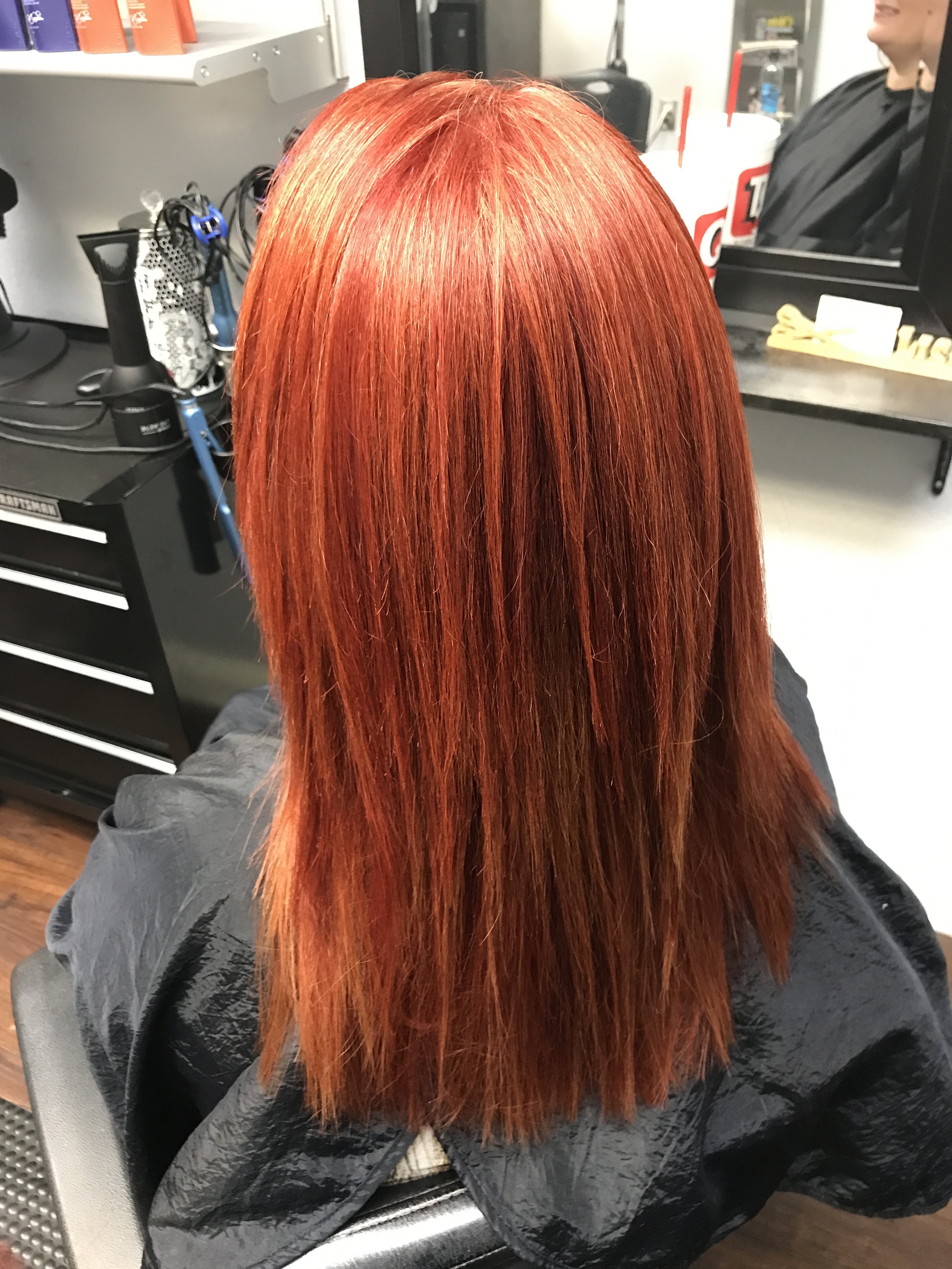 A person with long, straight, vibrant red hair sitting in a salon chair, wearing a black cape.