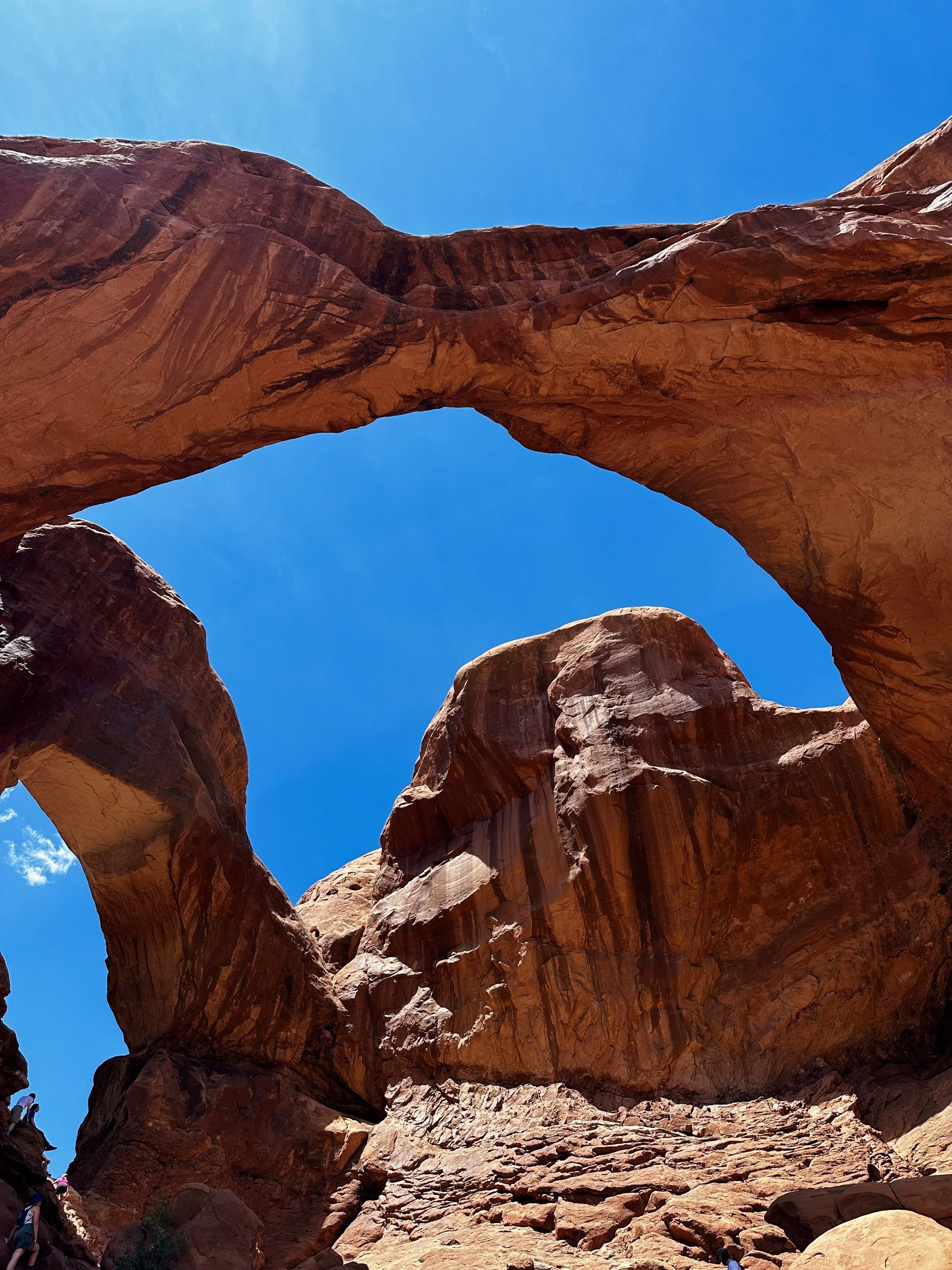 A large natural rock formation with a prominent arch set against a bright blue sky. The rocks are reddish-brown with textured surfaces, and there are people visible at the bottom for scale.