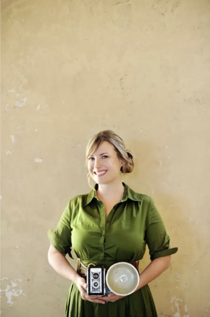 A woman in a green dress holding an old-fashioned camera and a tray with a candle, standing against a plain beige wall.