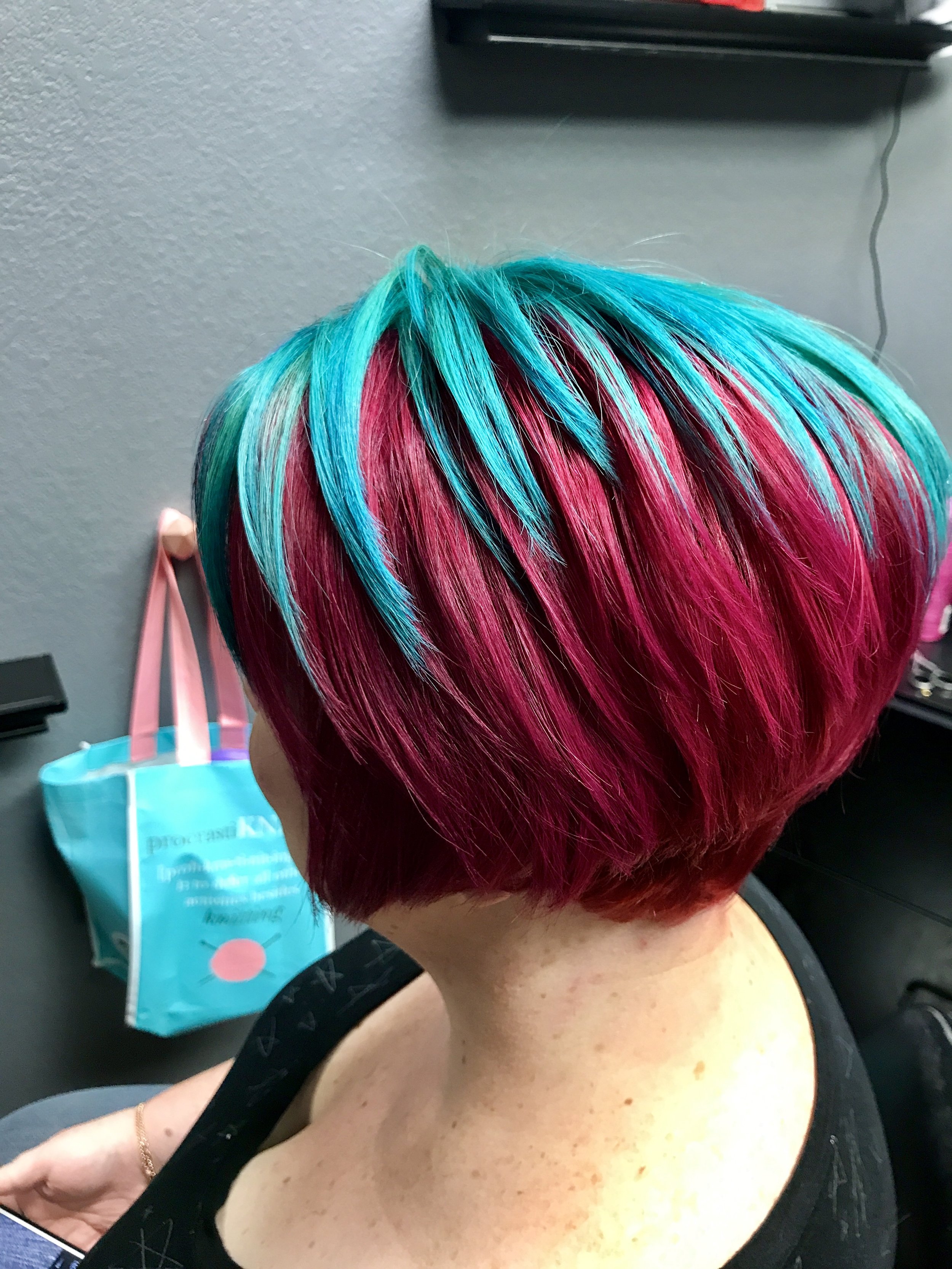 A person with short, vibrant, multi-colored hair that is teal at the top and pink at the bottom, with a rounded bob style. The person is wearing a black top with a wide neckline, revealing their shoulders and upper chest. The background shows a gray wall, a black shelf, and a blue and pink tote bag hanging on a hook.