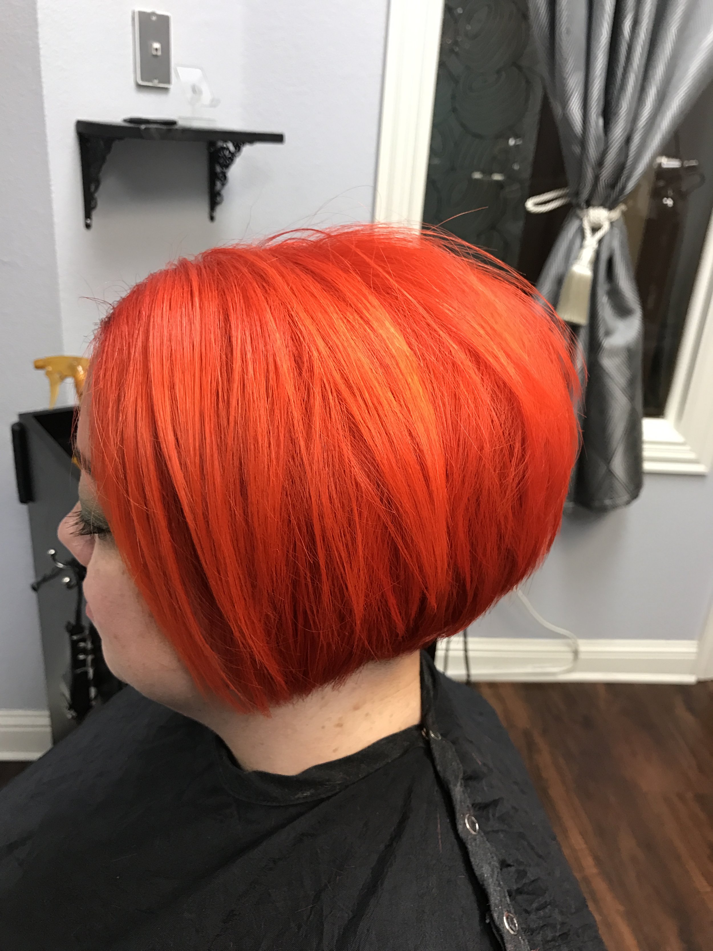 Side view of a person with bright red hair in a short bob haircut, wearing a black hoodie, in a domestic or salon setting.
