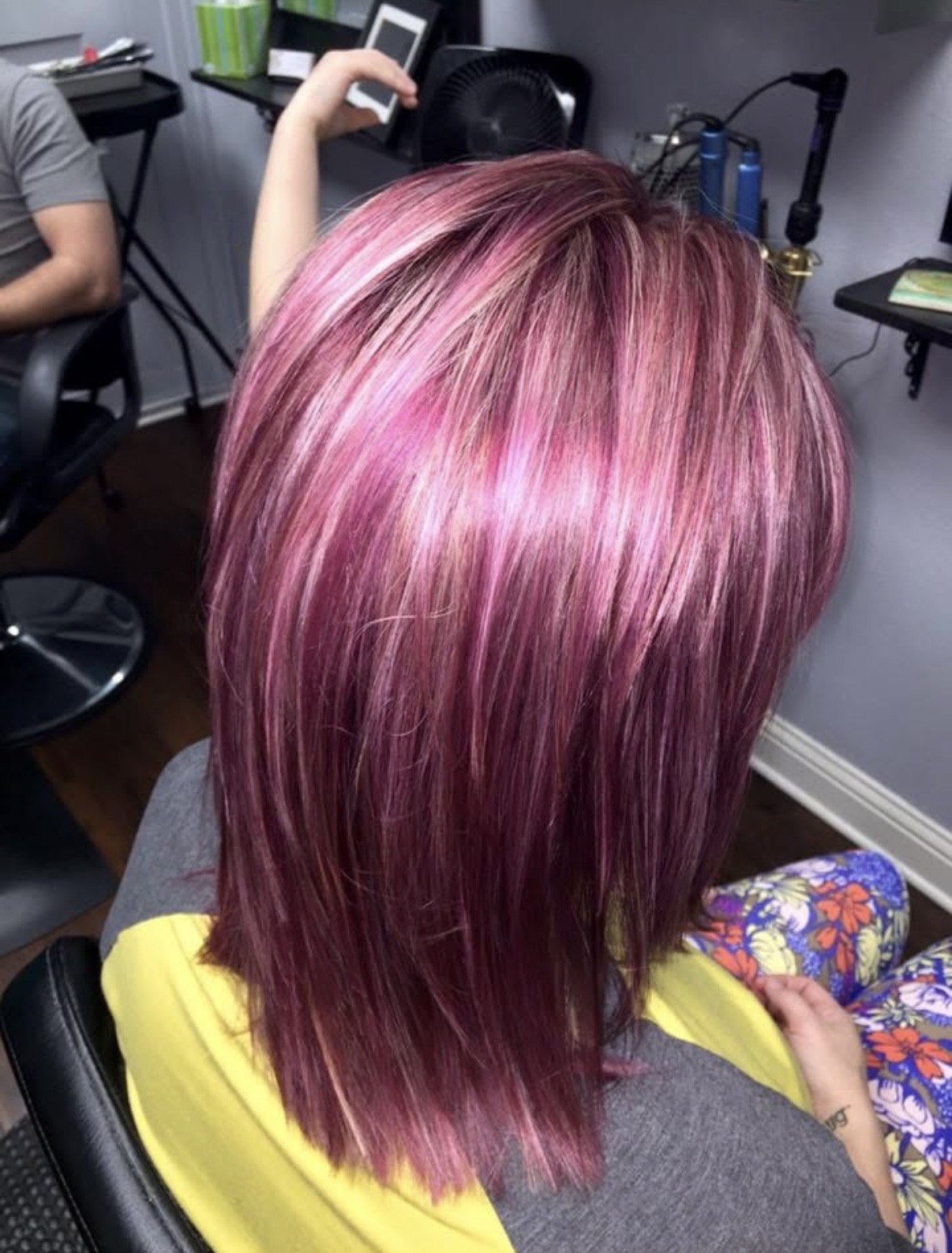 Person with pink, purple, and blonde hair sitting in a salon chair, with the hairstylist visible in the background.