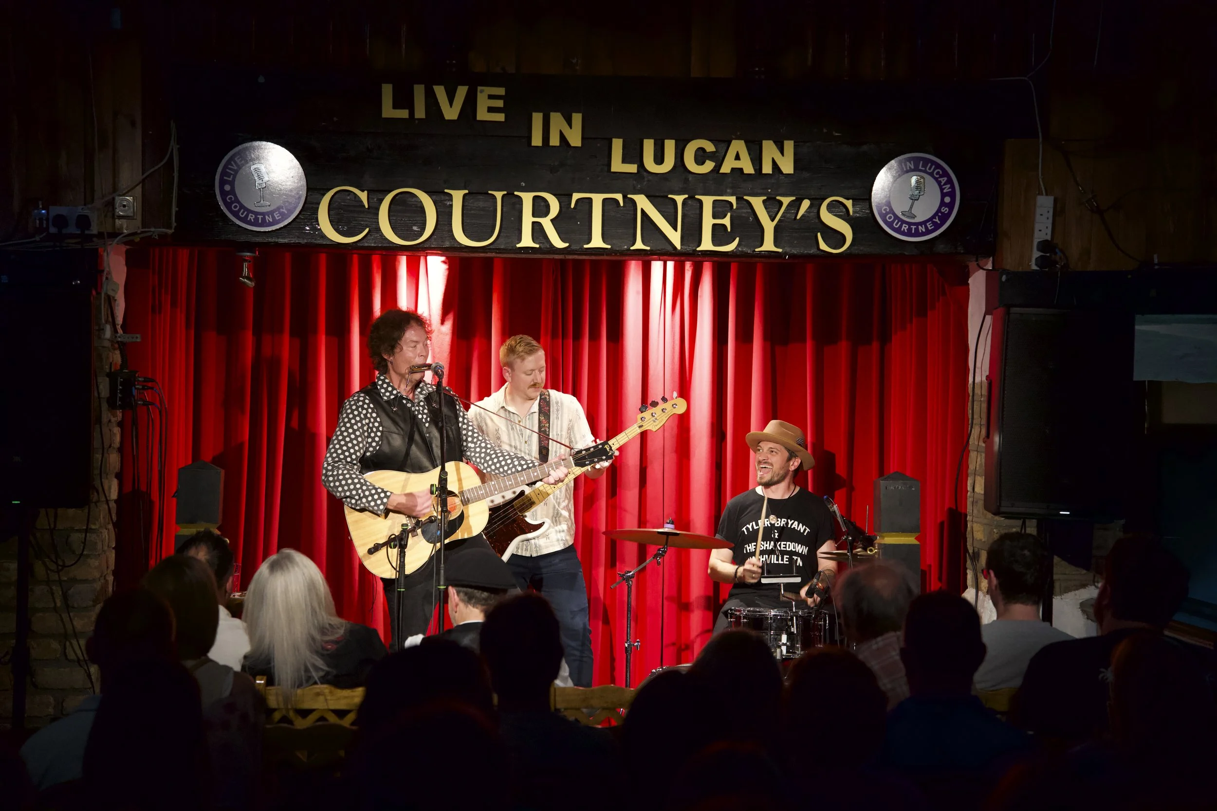 Musicians performing on stage at live music venue with red curtains and sign reading "Live in Lucan Courtney's".