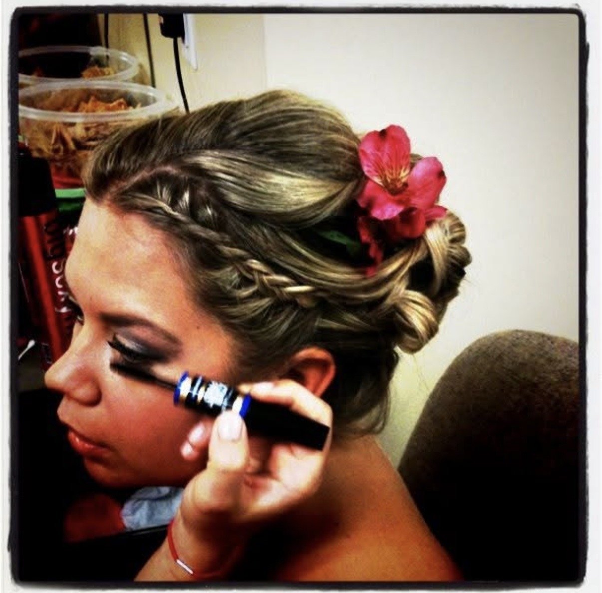 A woman with styled blonde hair adorned with a large red flower, having eye makeup applied with a black eyeliner pencil.