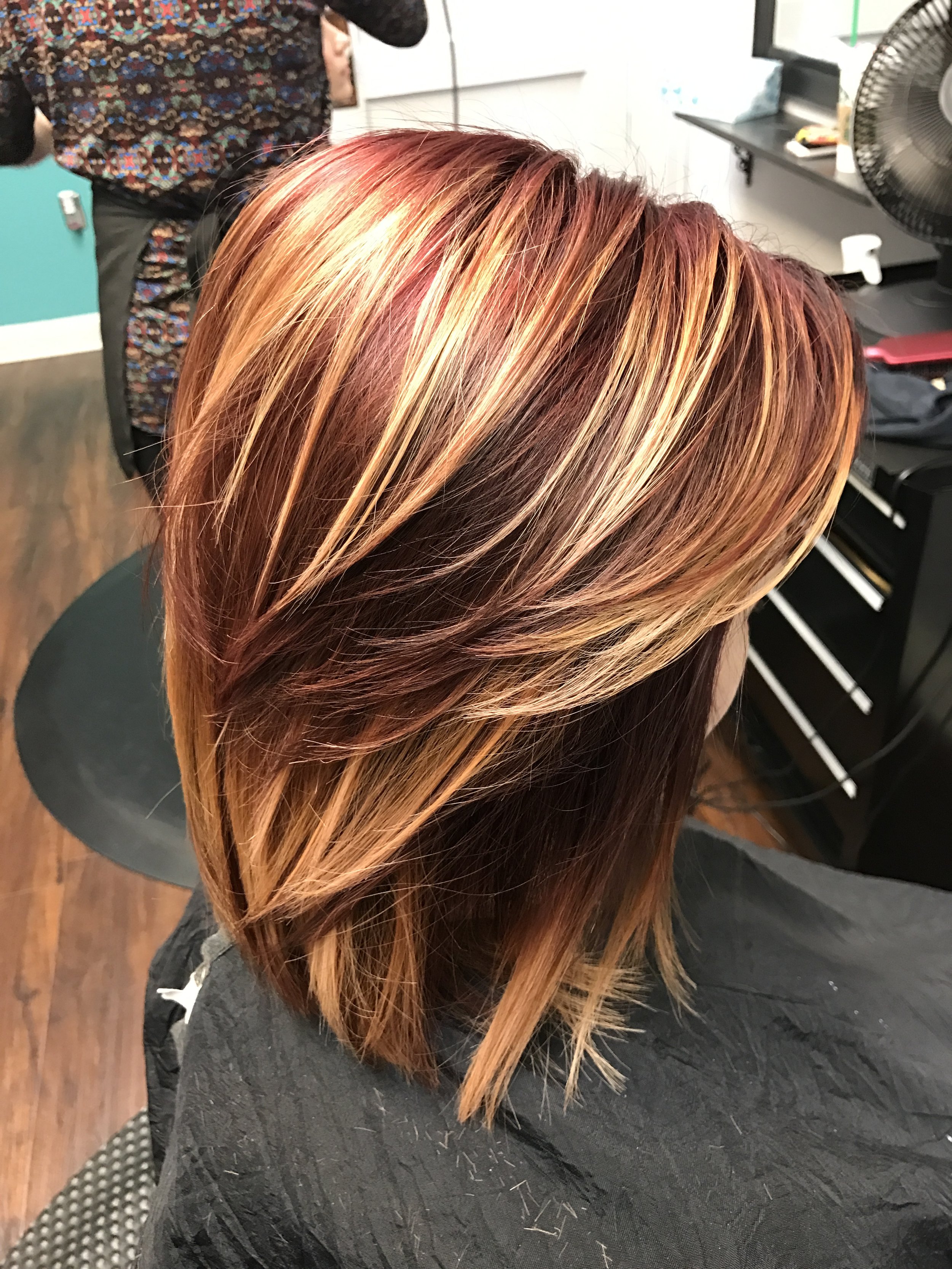 Close-up of a person's hair styled with caramel and auburn highlights, parted to the side, with a salon environment in the background.