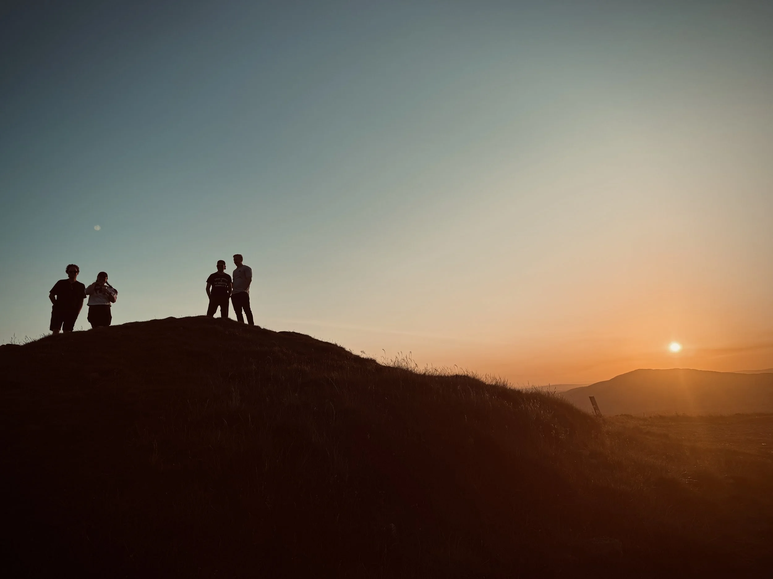 Four people standing on a grassy hill during sunset, with the sky transitioning from blue to orange and the moon visible.