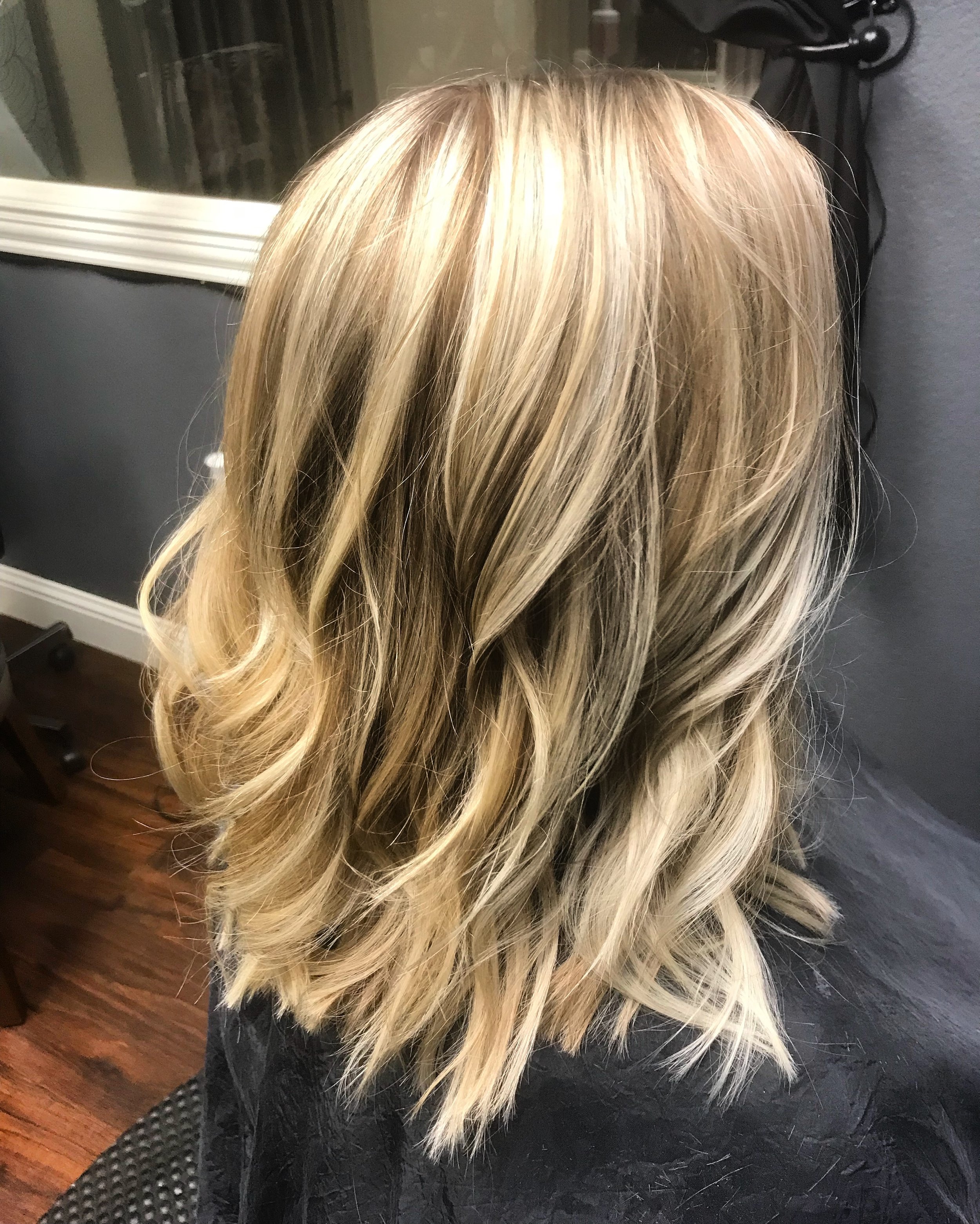 Blonde hair with loose curls resting on a black salon cape, seen from the back in a salon setting with a window and furniture in the background.