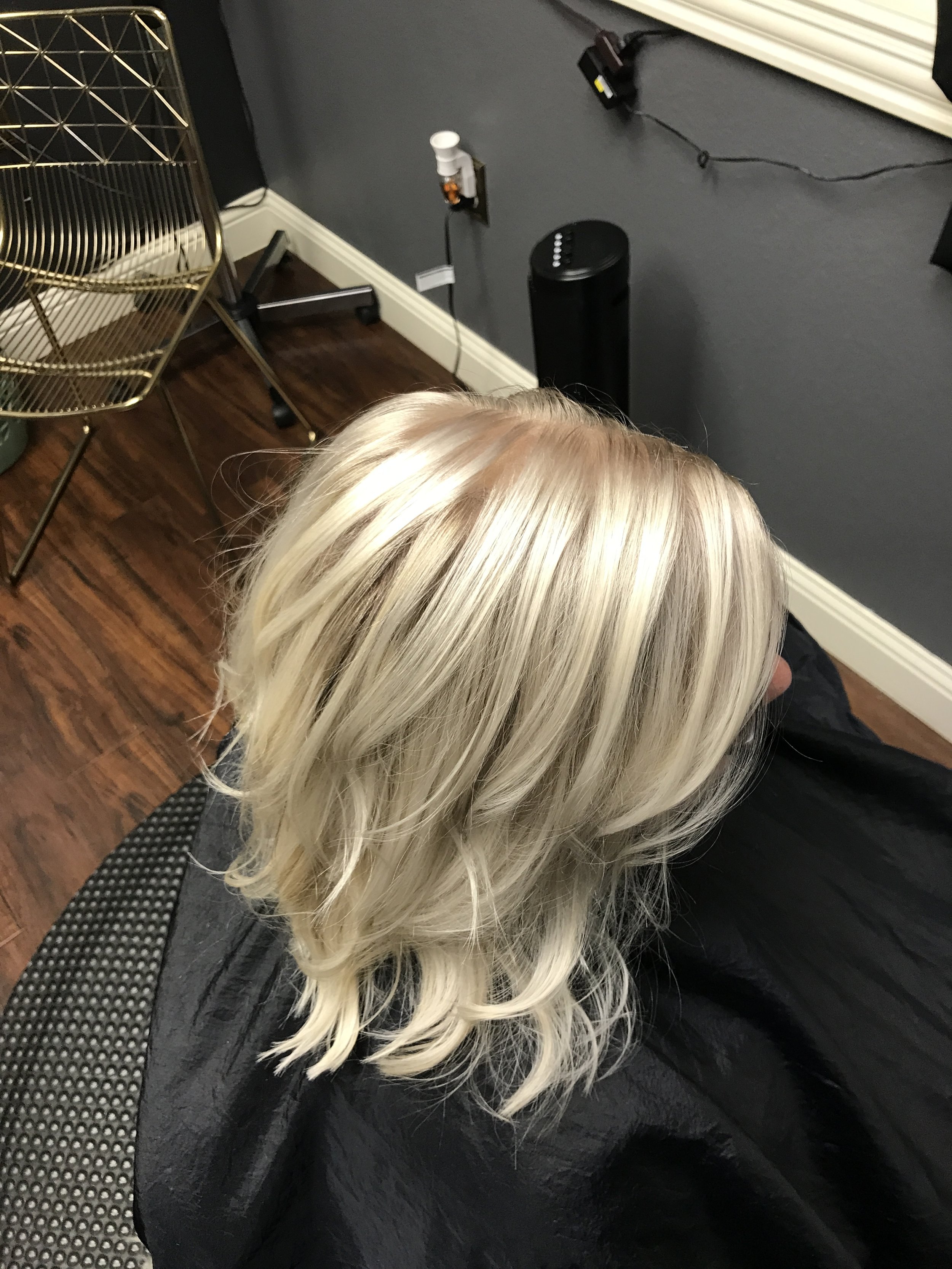 Side view of a person with long platinum blonde hair sitting in a salon chair, with a black cape around their shoulders. In the background, there's a chrome hairdryer hood, a gold metal chair, and salon equipment on the dark gray wall.