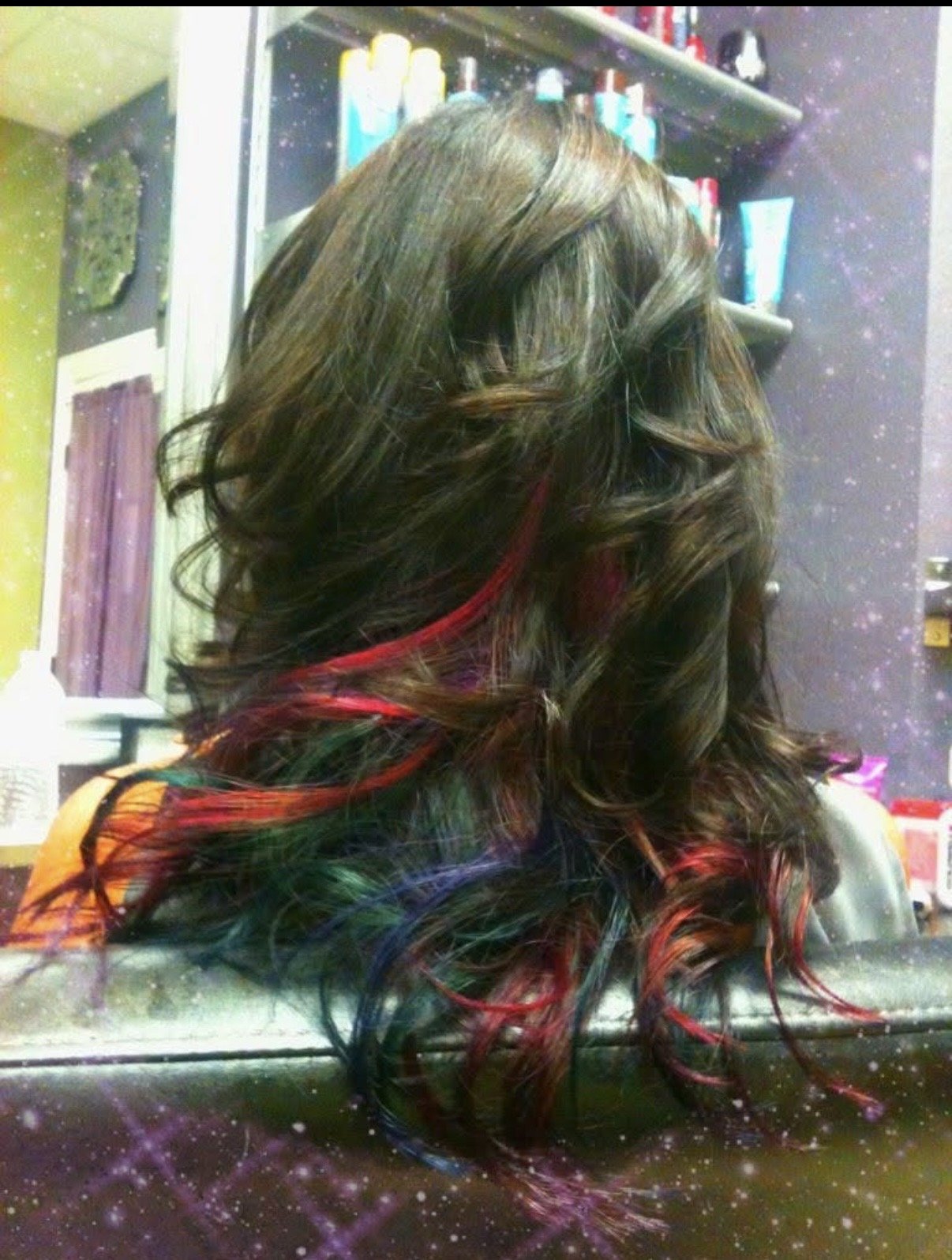 A person with long, wavy hair that has vibrant red, green, and blue streaks, sitting in front of a mirror at a salon or hairdresser shop.