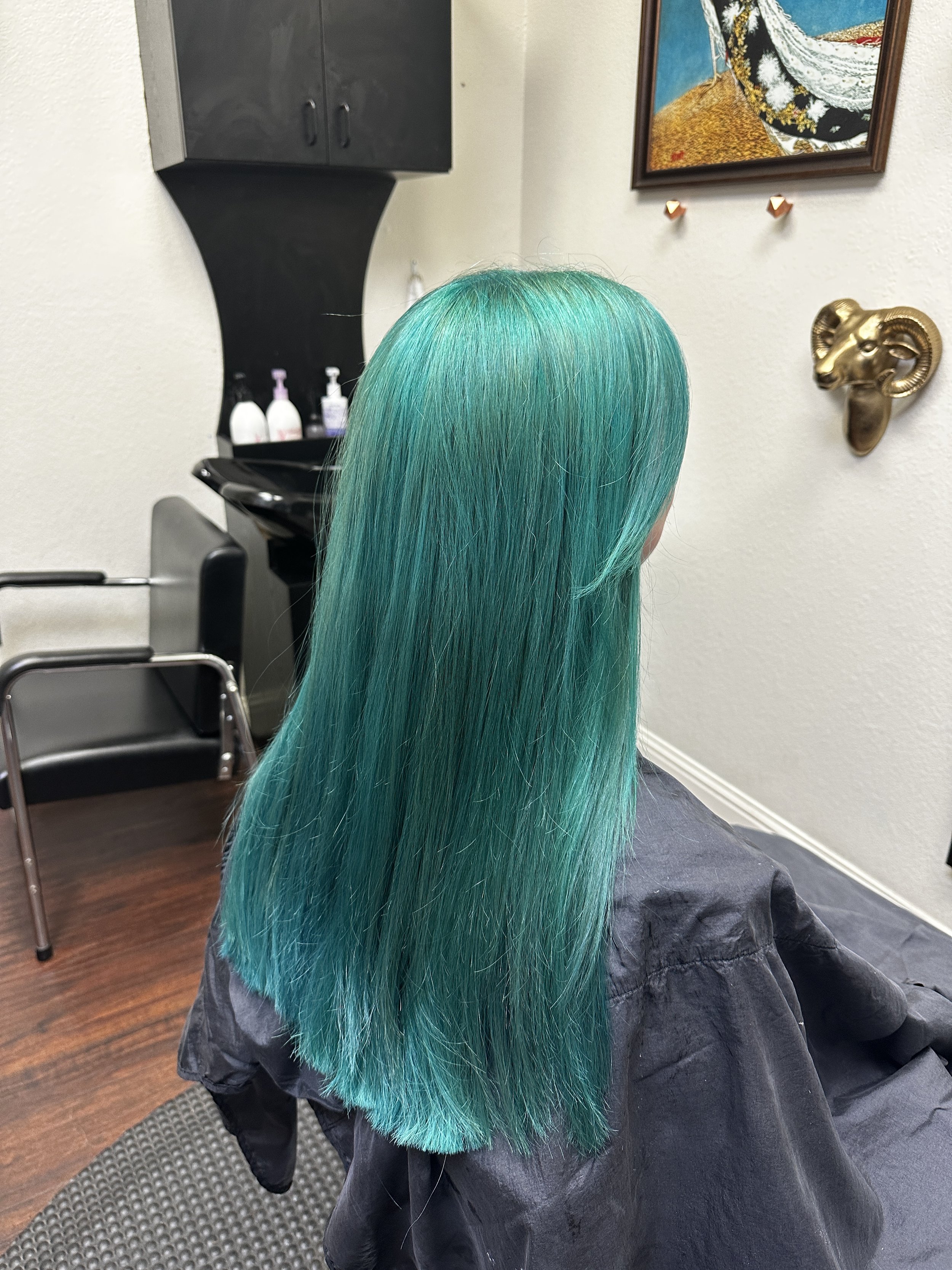 Back view of a person with long, straight, vibrant teal hair sitting in a salon chair, wearing a black cape, in a hair salon with wall art, a clock with ram horns, and a mirror in the background.