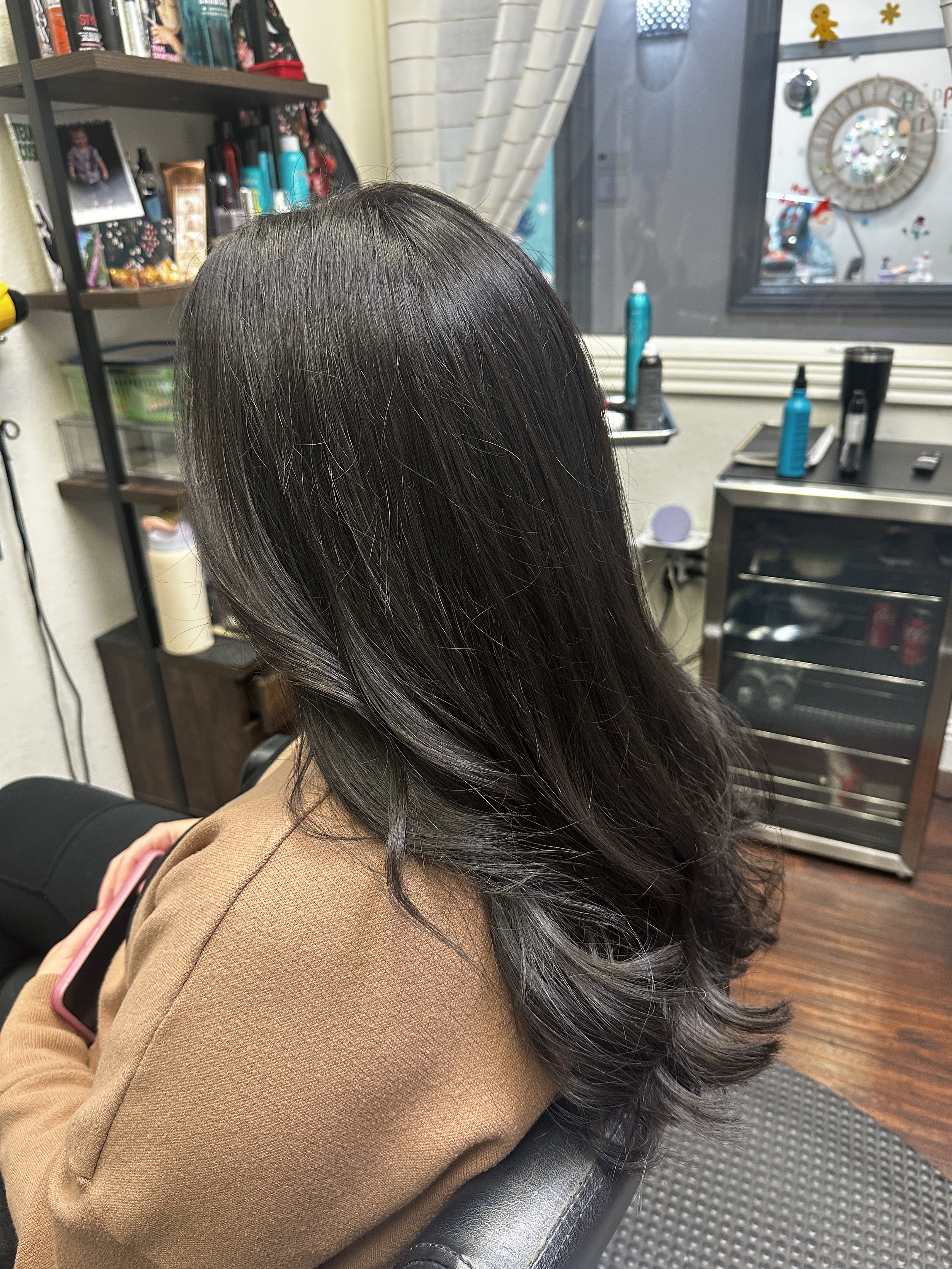 A woman with long, dark, shiny hair sitting in a salon chair. She is wearing a tan blazer and is holding a pink smartphone. The background shows salon shelves with hair products, a window with decorations, and a small metallic storage cart.
