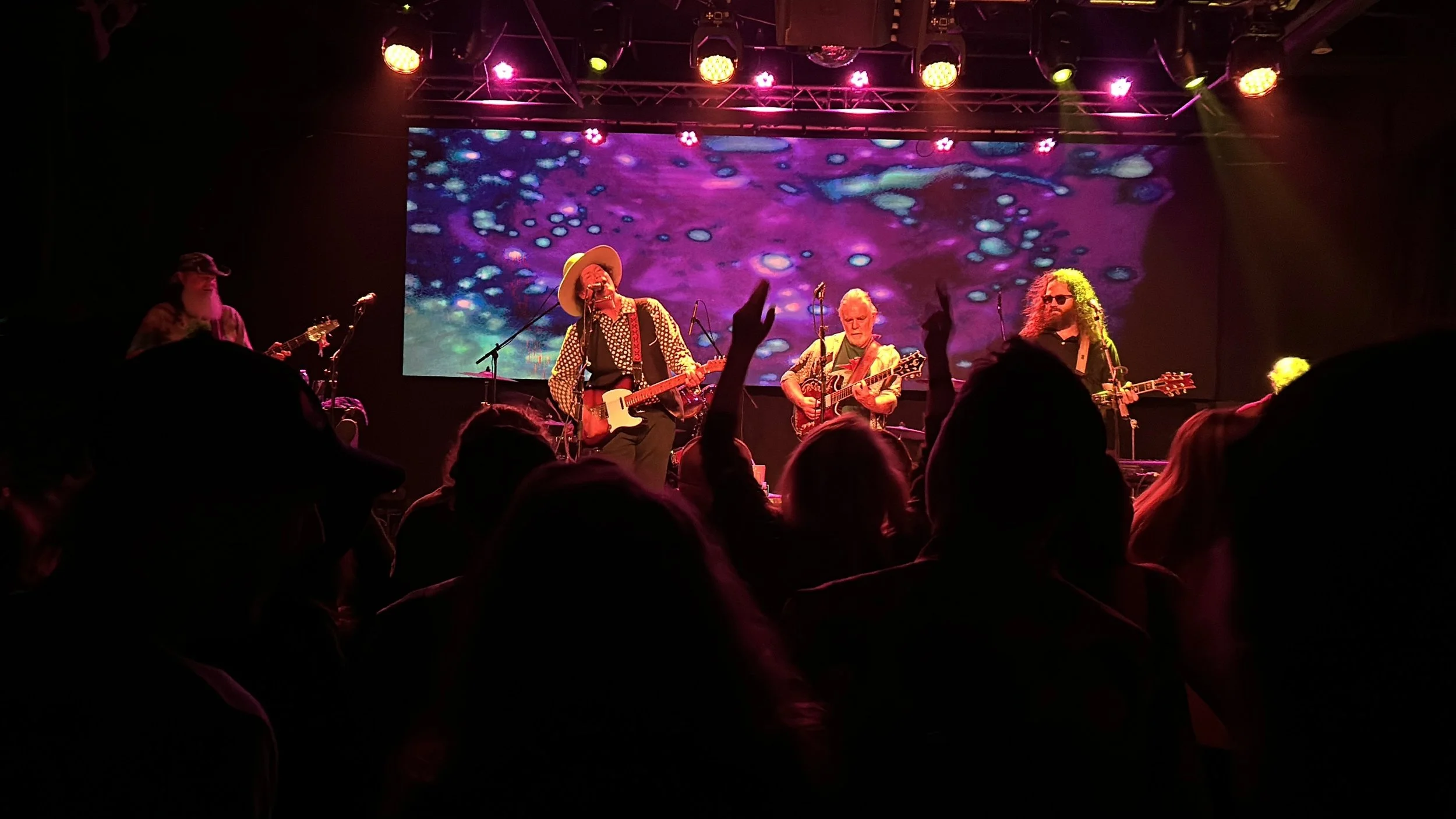 A band performing live on stage with four musicians, colorful visual effects on a large screen behind them, and audience members visible in the foreground.