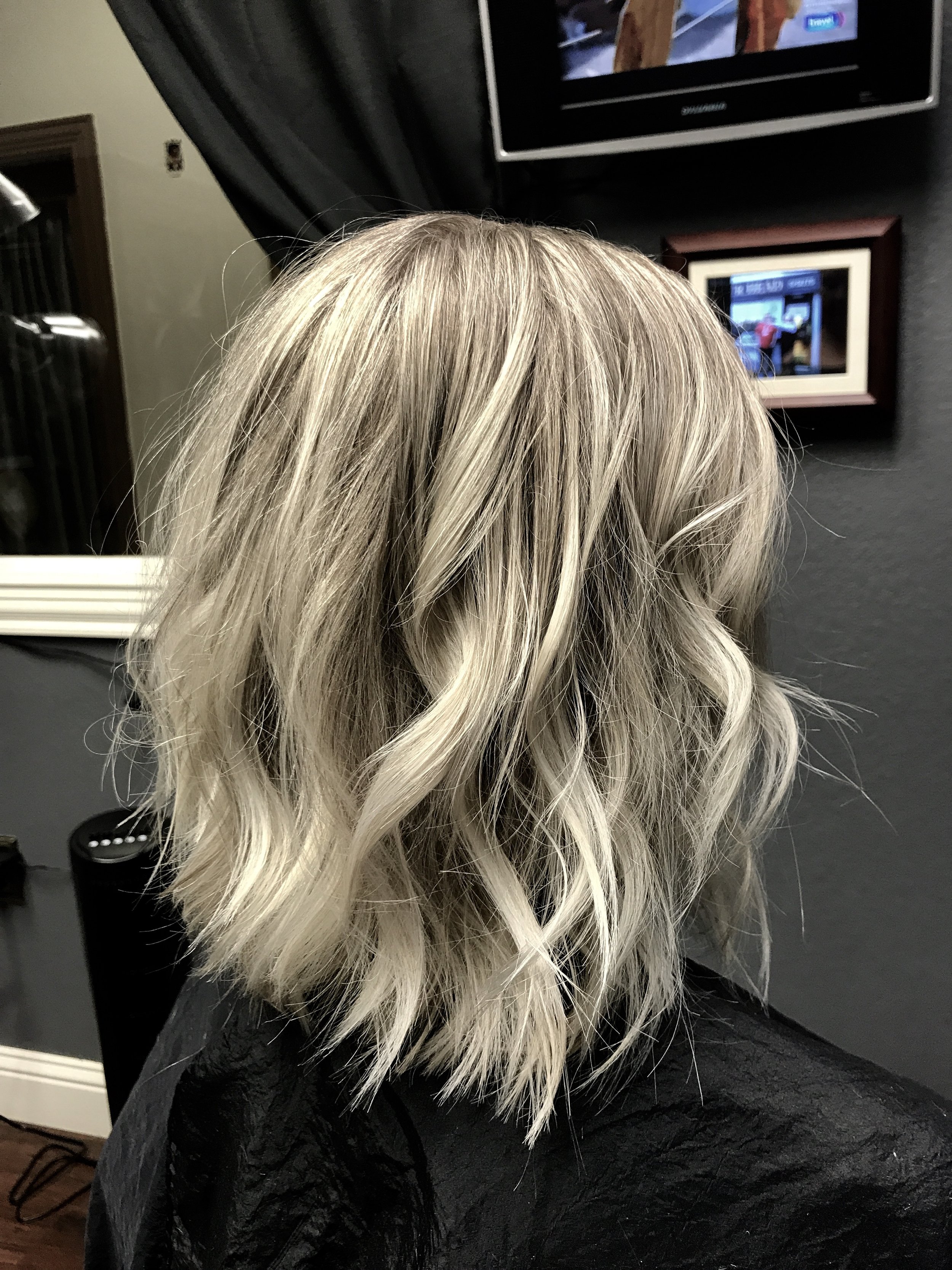 Woman with wavy blonde hair sitting in a salon chair, facing away, with a mirror and television in the background.