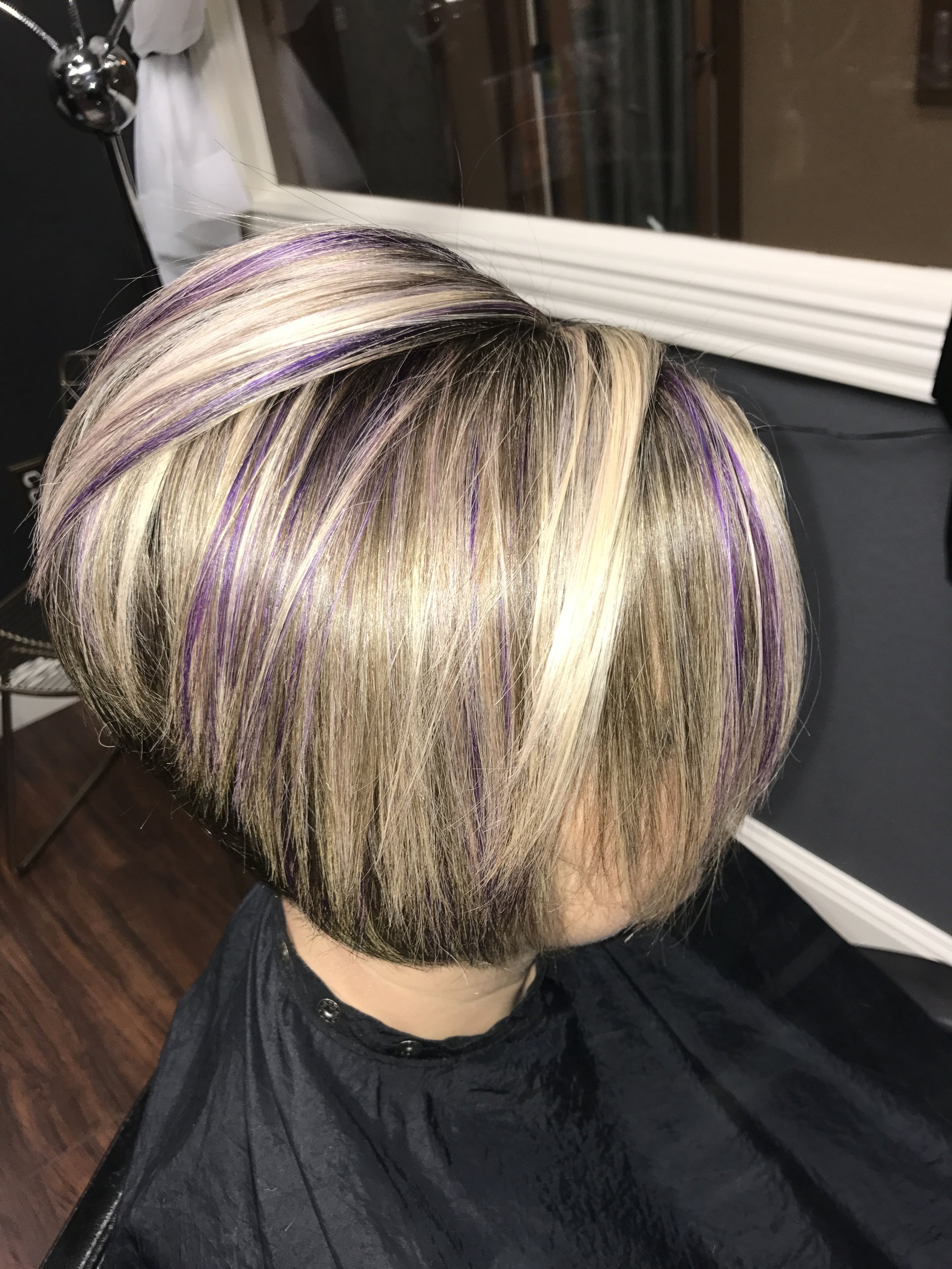 A person with blonde hair featuring purple highlights at the roots, side-swept bangs, in a salon chair wearing a black cape.