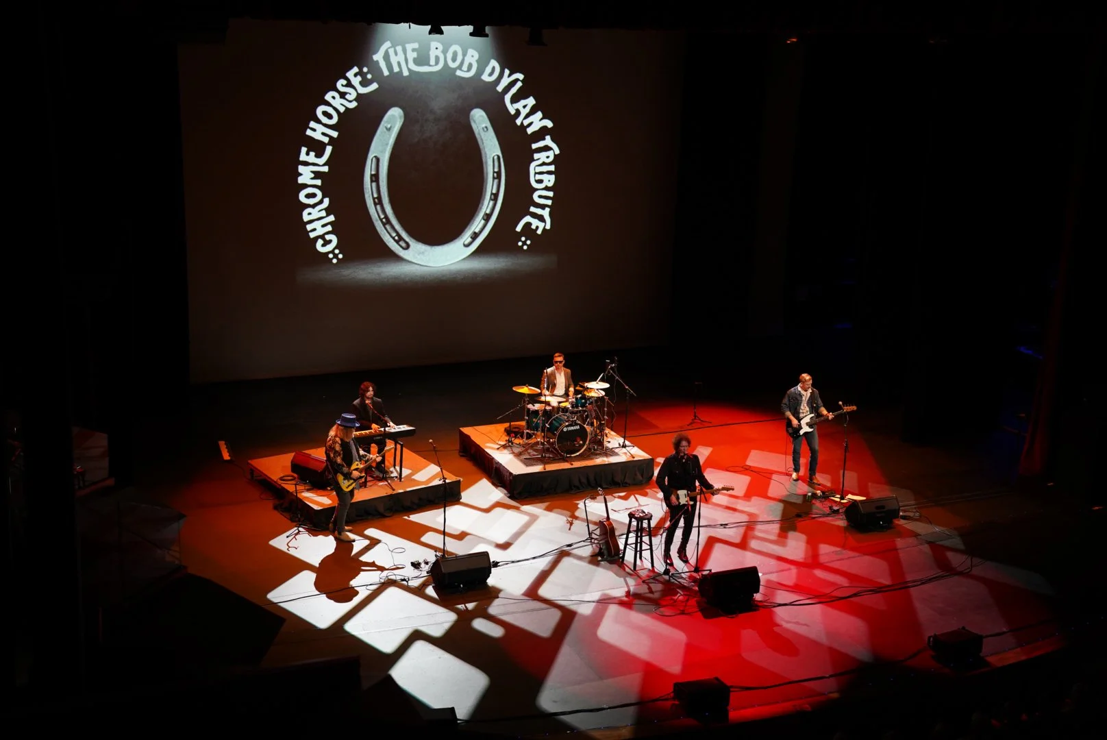 A band performing on stage with a large screen behind them showing the band name 'The Bob Dylan Ruge'. There are five members: one playing keyboard, one on drums, two with guitars, and one singing.