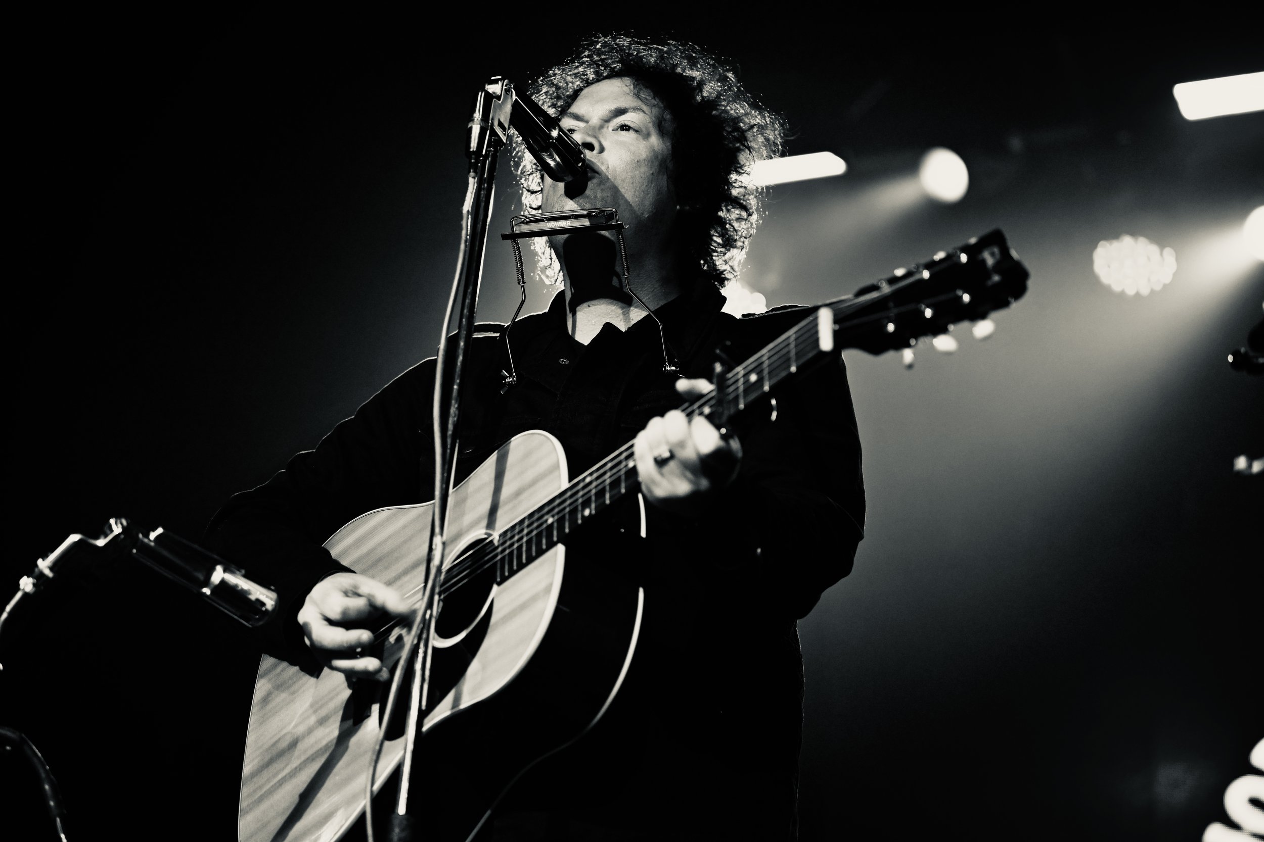 Black and white photo of a male musician playing an acoustic guitar and singing into a microphone on stage with bright stage lights in the background.