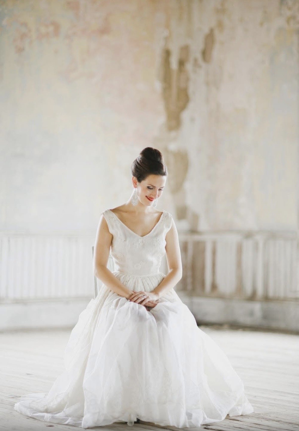 A woman in a white sleeveless wedding dress sitting in a room with a textured wall and wooden floor, her hair in an updo, smiling and looking downward.