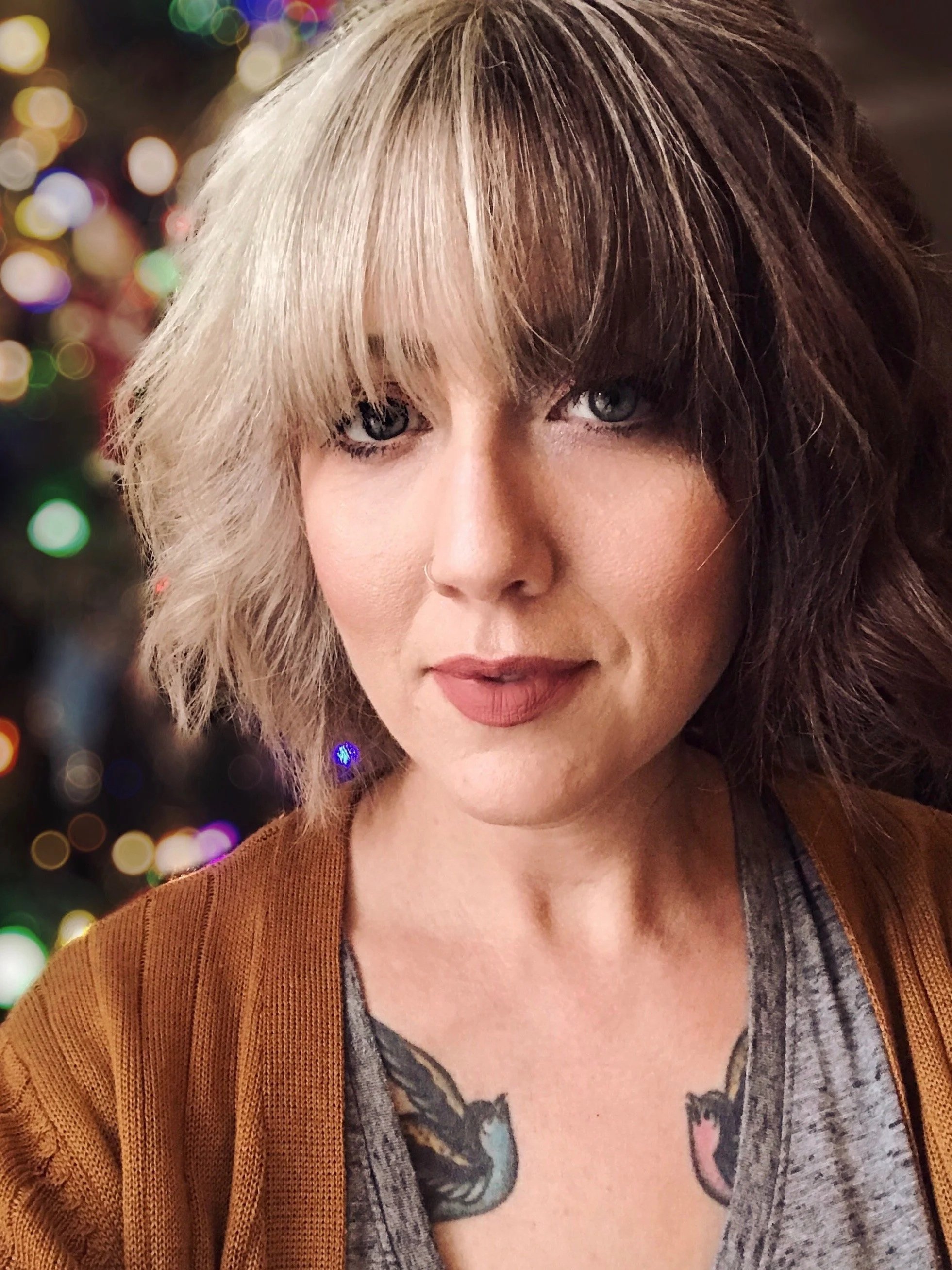 A woman with shoulder-length blonde hair and bangs, wearing a brown jacket and a gray shirt, with tattoos visible on her chest, and blurred colorful lights in the background.