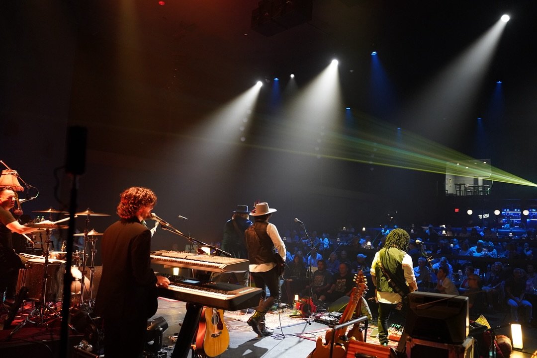 A band performing on stage with musical instruments, including keyboards, guitars, and drums, under stage lights in front of an audience.